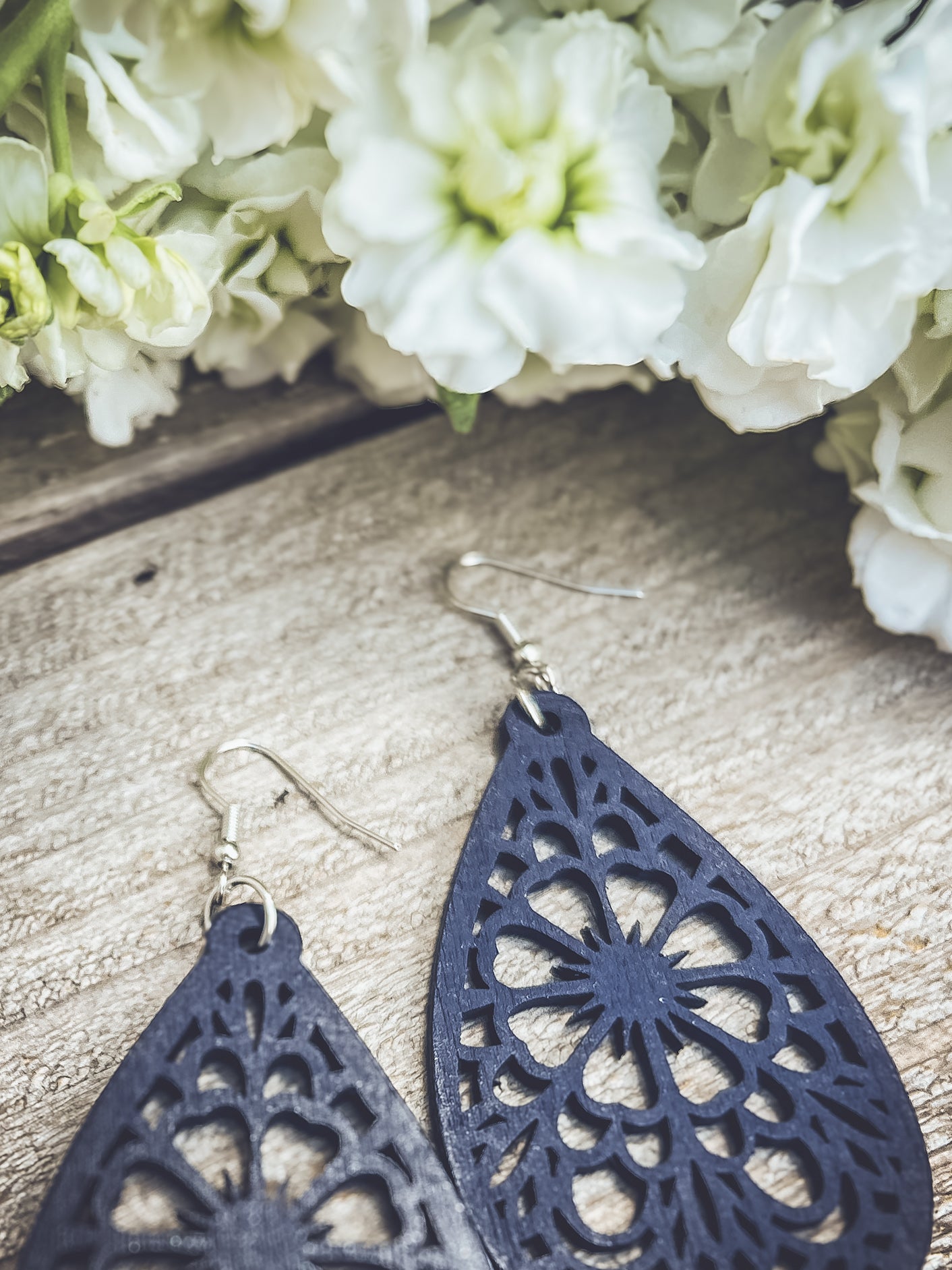 Photography of dark blue teardrop wooden earrings featuring intricate laser-cut floral patterns and silver hooks, resting on a rustic wood surface with a soft-focus cluster of white stock flowers displaying pale green centers
