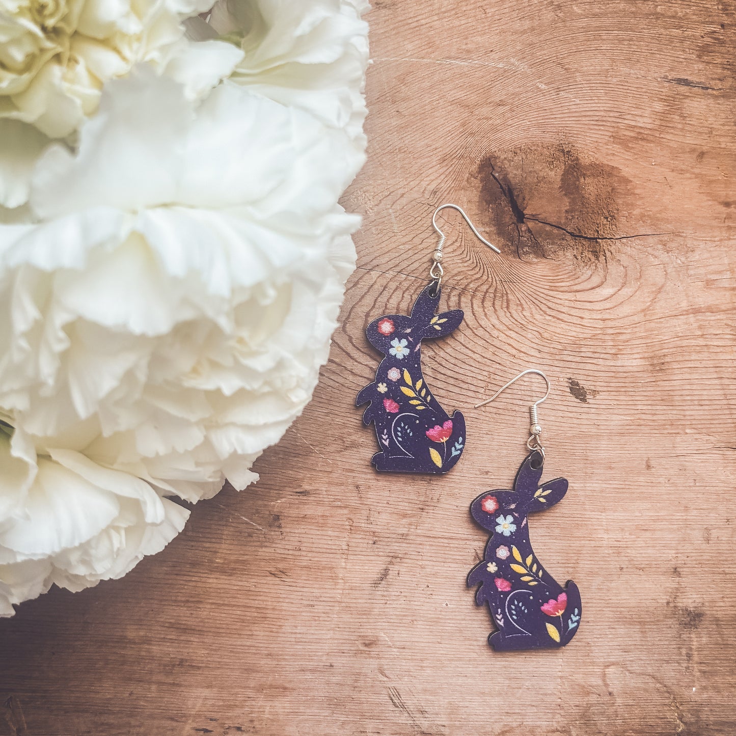 Pair of wood rabbit earrings on a rustic wooden surface next to fluffy white flowers featuring dark blue bunnies with vibrant pink, yellow, and light blue floral patterns and silver hooks