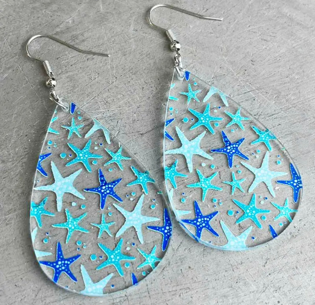 Clear acrylic teardrop earrings patterned with vibrant blue and light blue starfish accents, complemented by tiny blue dots and silver fishhook ear wires