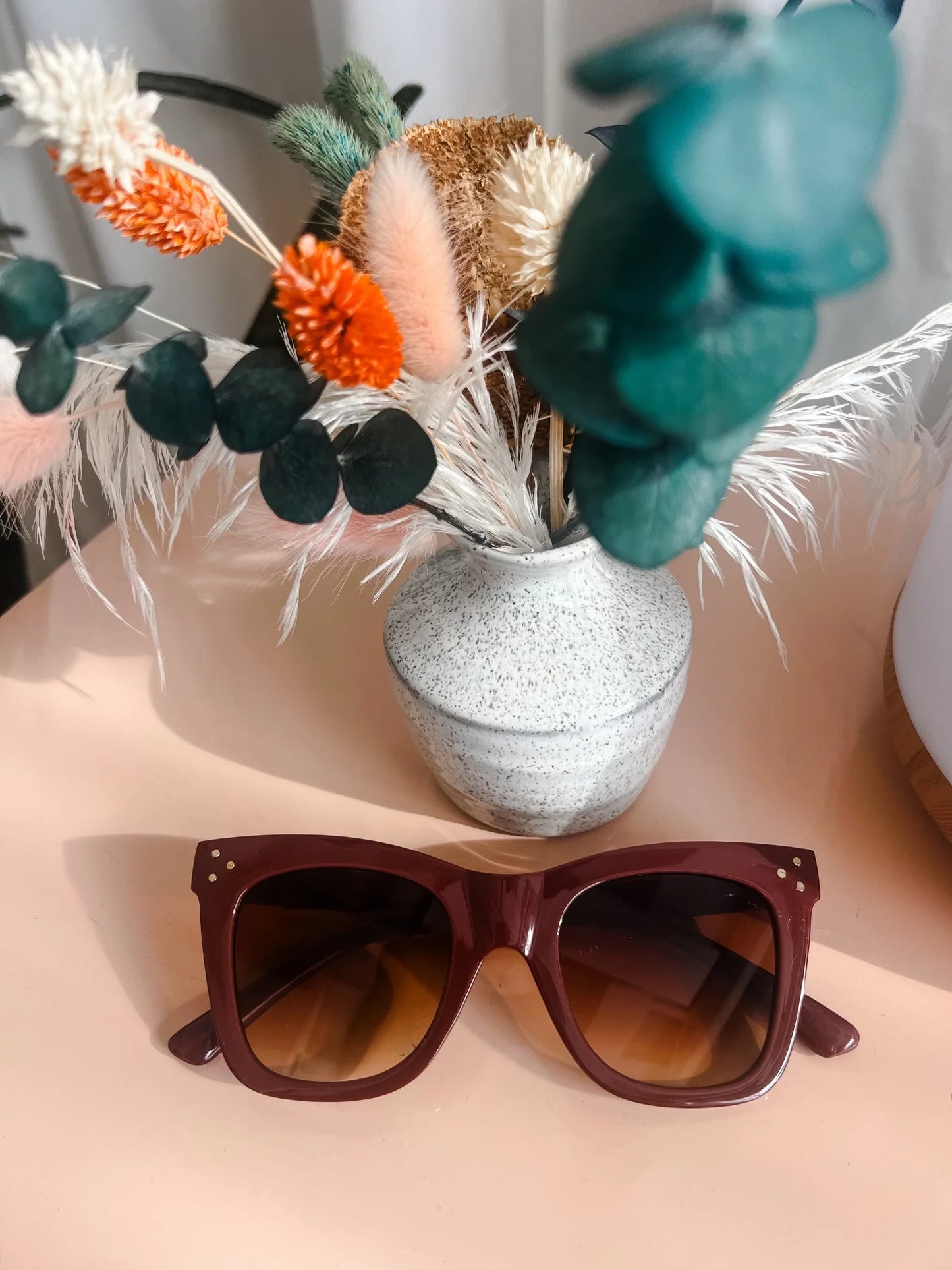 A still life photograph of a speckled grey ceramic vase filled with colorful fluffy dried flowers including orange, white, pink, and dark green eucalyptus, beside burgundy sunglasses with brown gradient lenses and subtle gold stud accents, illuminated by sunlight on a peach tabletop