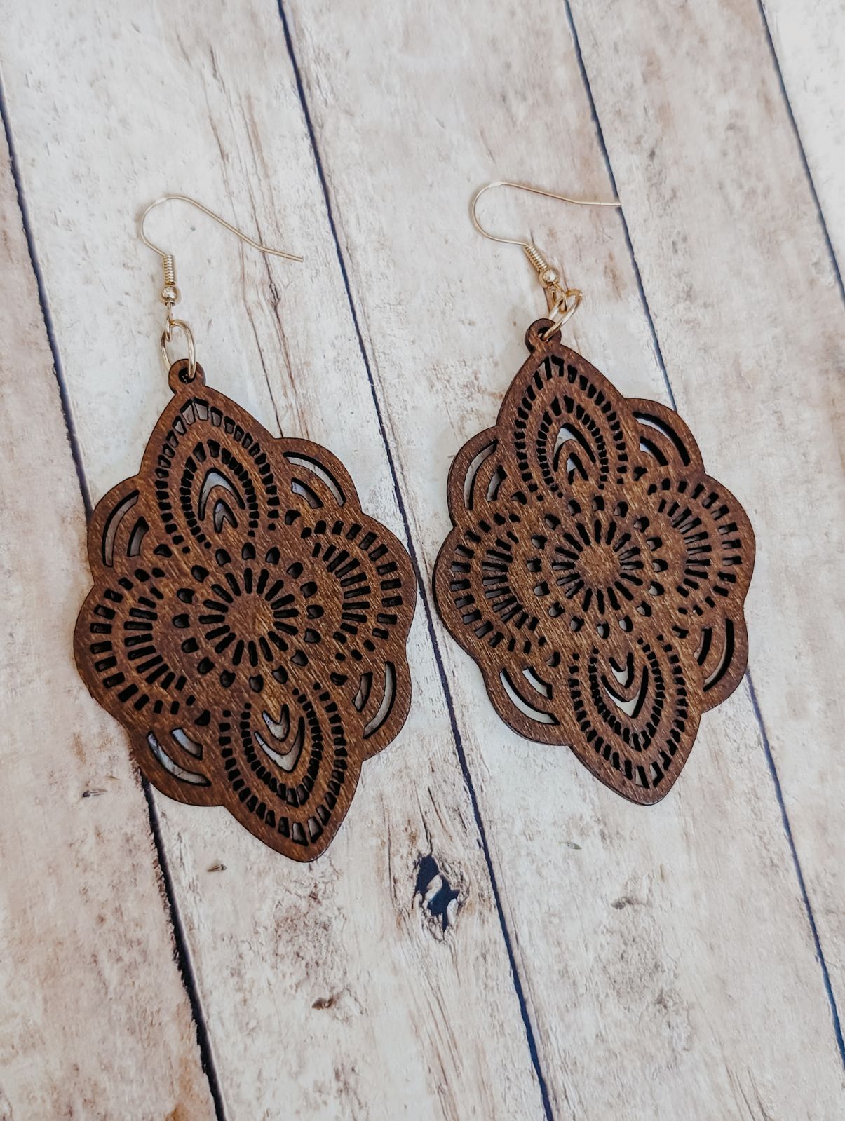 Wooden earrings featuring dark brown laser-cut quatrefoil shapes with intricate mandala patterns and silver fish hook ear wires displayed on a light wood grain background