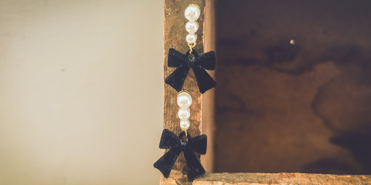 A pair of dangling earrings featuring three white pearls linked by gold accents and a black velvet bow, suspended from a textured wooden beam
