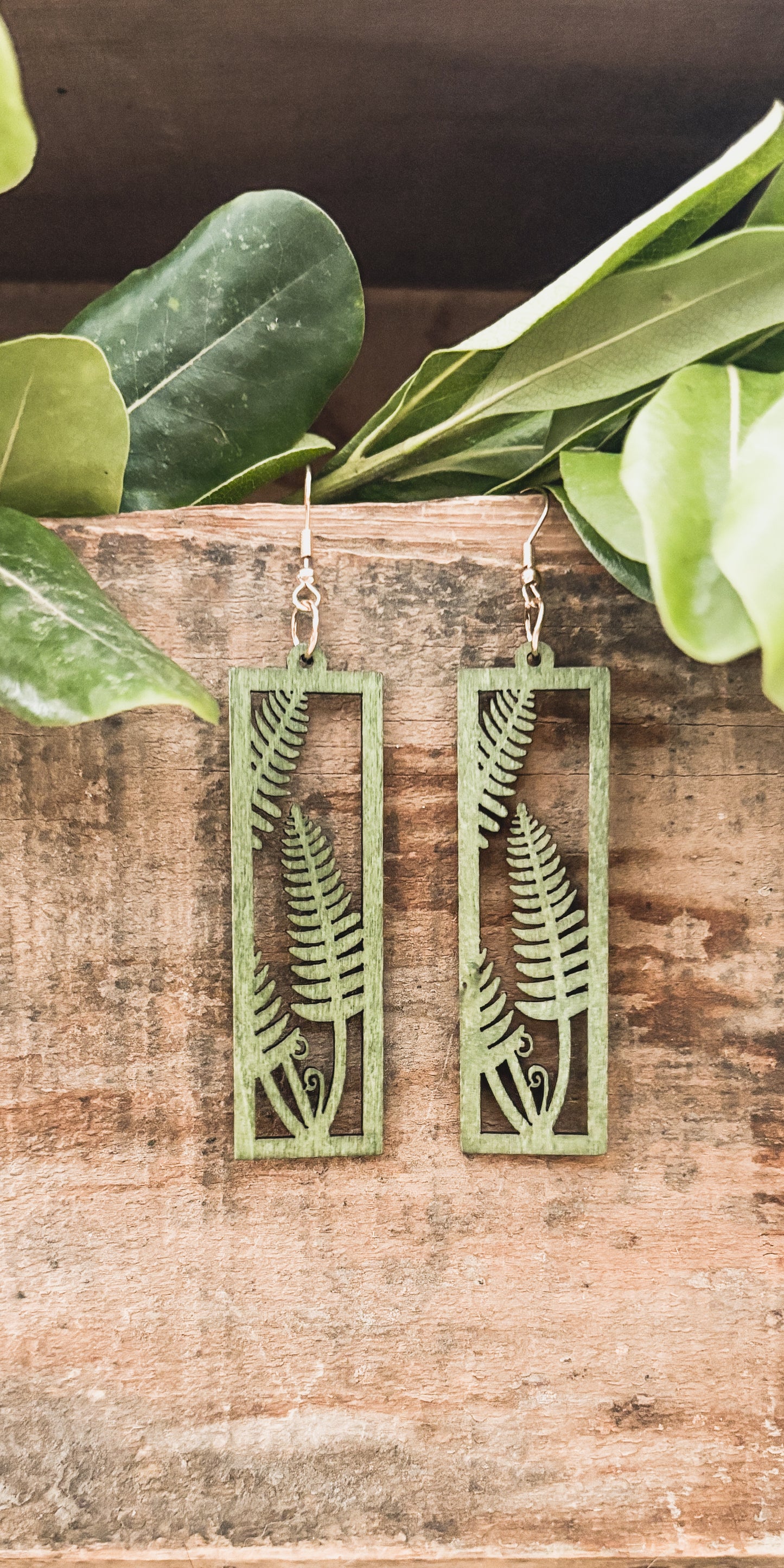 Product photography of a pair of light green rectangular wooden dangle earrings featuring intricate fern cutouts and gold-colored hooks displayed against a rustic wooden surface accented by vibrant green leaves