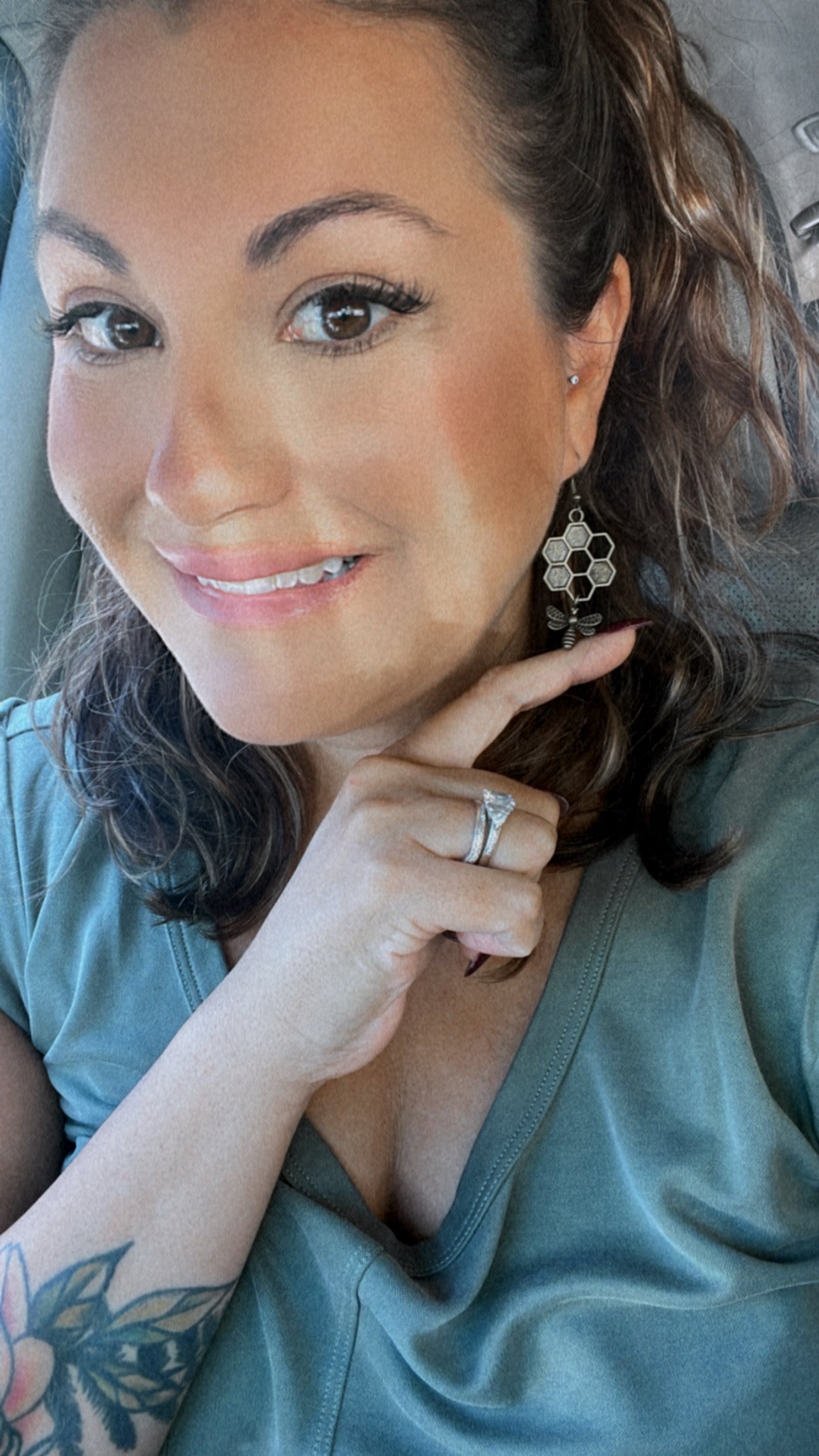 A smiling woman in a car pointing to a hexagonal honeycomb and bee earring wearing a large diamond engagement ring and a floral arm tattoo