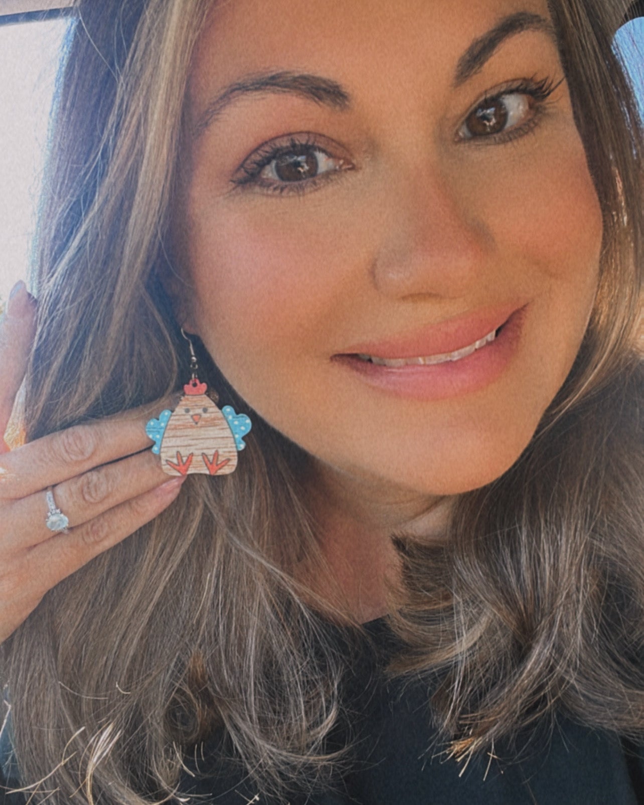 Photograph of a smiling woman with brown hair displaying a wooden chicken earring featuring blue polka dot wings red comb and orange feet, wearing a diamond engagement ring