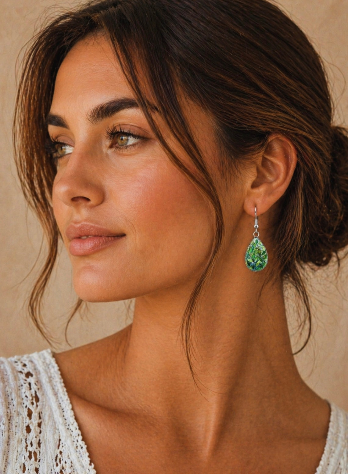 Close-up photograph of a woman with a warm tan complexion and expressive amber eyes, her dark hair styled in a loose bun with a few wispy strands framing her face, wearing a white lace top and a vibrant green teardrop-shaped earring intricately detailed with a leaf pattern
