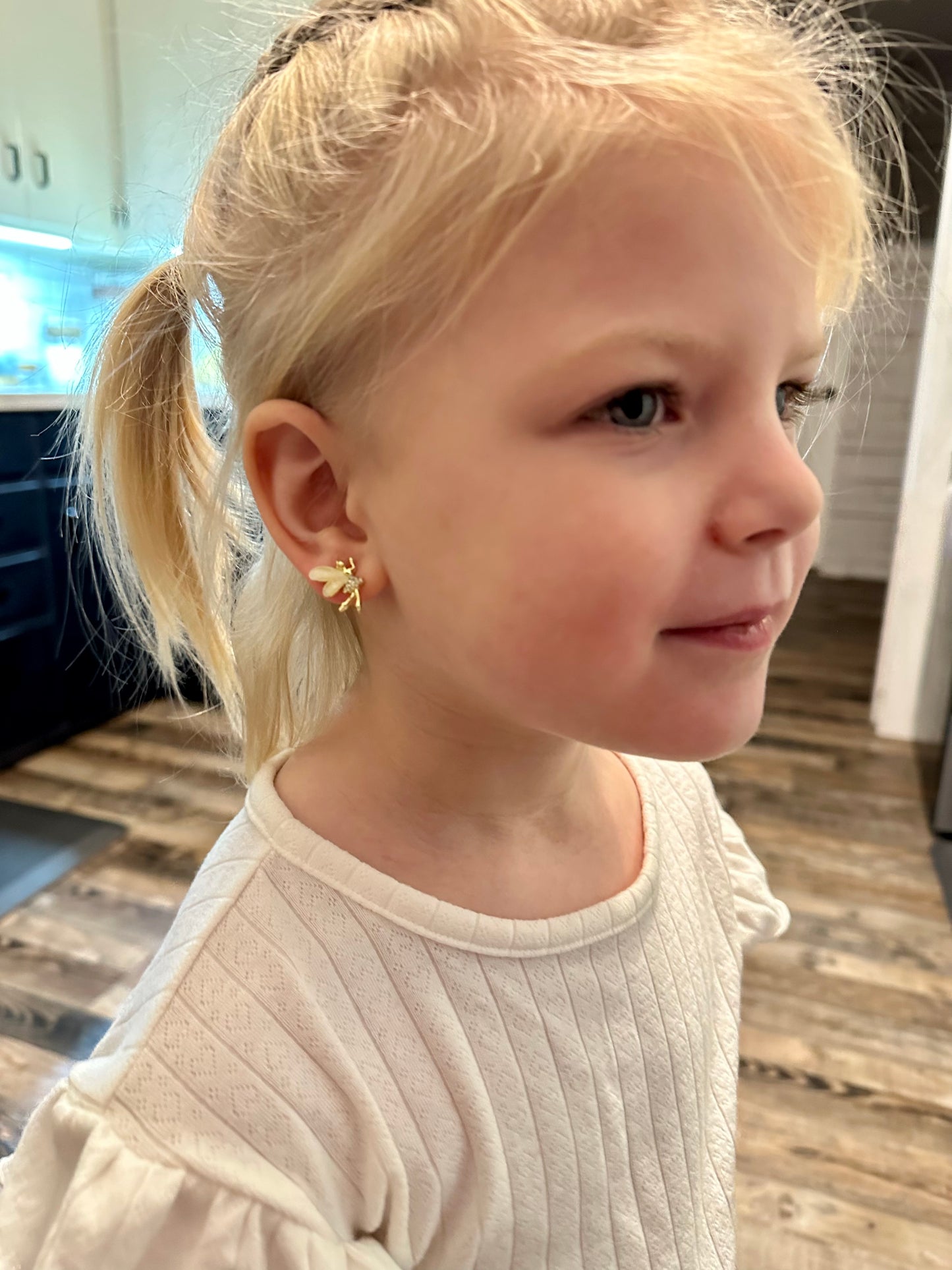 Photograph of a young girl with blonde hair in pigtails wearing a gold and white bee earring and a white textured shirt indoors