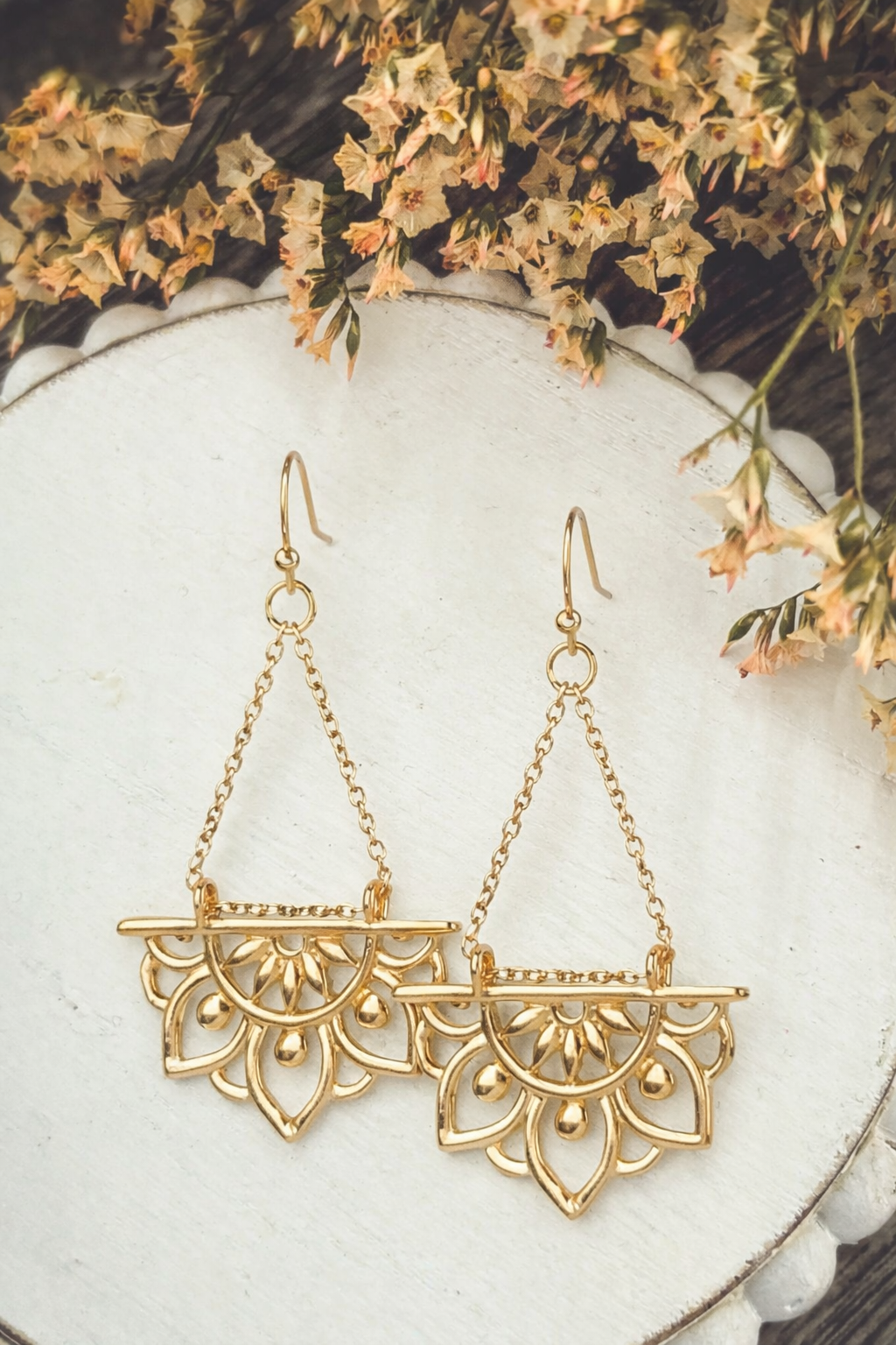Gold-toned dangle earrings featuring a semi-circular mandala-inspired design with delicate chain drops displayed on a white scalloped-edge surface against a blurred background of dried cream and pale orange flowers