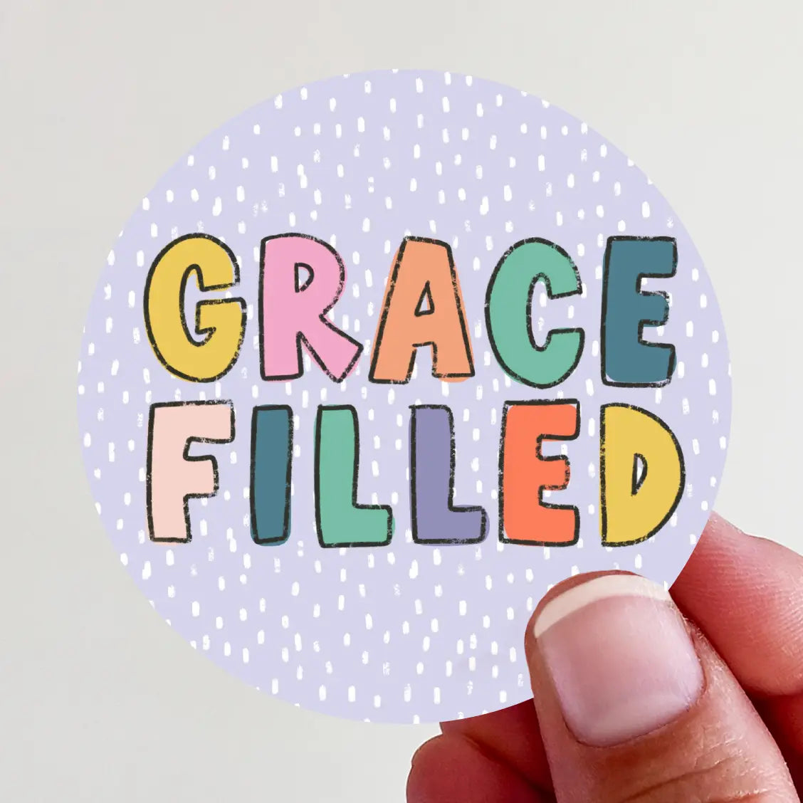 A circular sticker held by a person's hand, featuring the words GRACE FILLED in vibrant, distressed block letters of yellow, pink, orange, and teal against a light purple speckled background