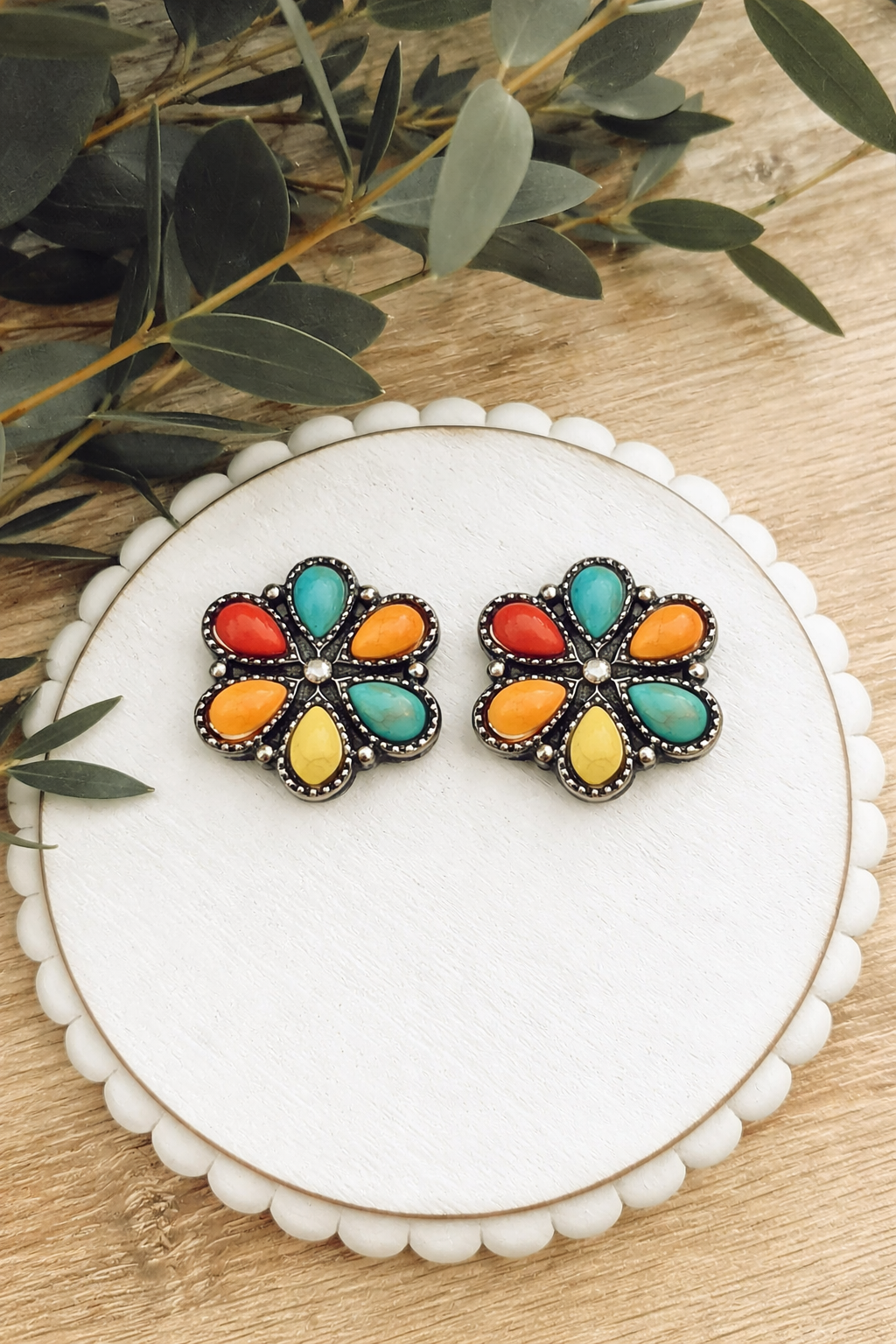A pair of vibrant flower-shaped earrings featuring red orange yellow and turquoise teardrop stones in a silver setting displayed on a white scalloped stand with eucalyptus leaves on a wooden background