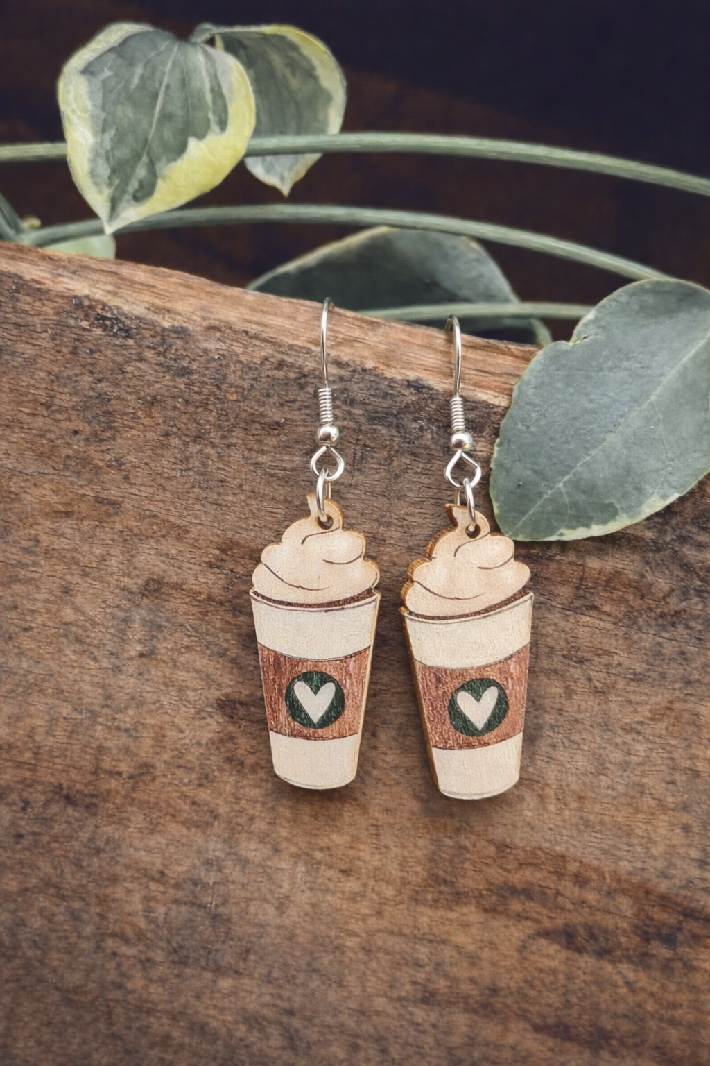 Handcrafted wooden coffee cup earrings with whipped cream tops and brown sleeves featuring green heart logos resting on a rustic wooden plank with variegated green leaves in the background