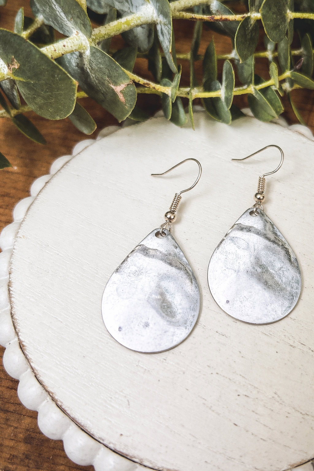 A pair of decorative teardrop earrings resting on a cream scalloped wooden coaster featuring a mottled silver finish simple fishhook wires and surrounded by green eucalyptus leaves on a rustic wood surface
