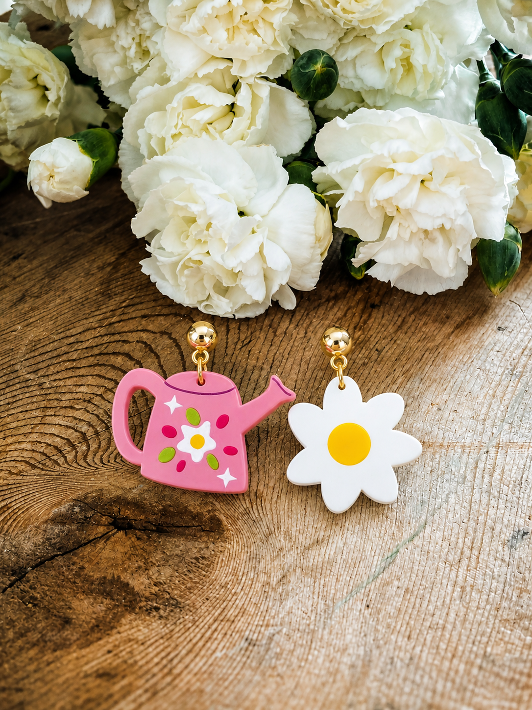 A pair of whimsical earrings featuring a pink watering can with a white flower and green leaf design alongside a white daisy with a yellow center, resting on a rustic wooden surface against a backdrop of soft white carnations