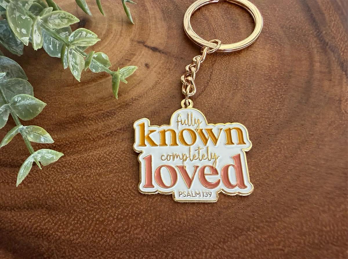 Digital photography of a white enamel keychain featuring the phrase "fully known completely loved PSALM 139" in orange and pink script on a textured brown wood surface accented by a green artificial plant sprig