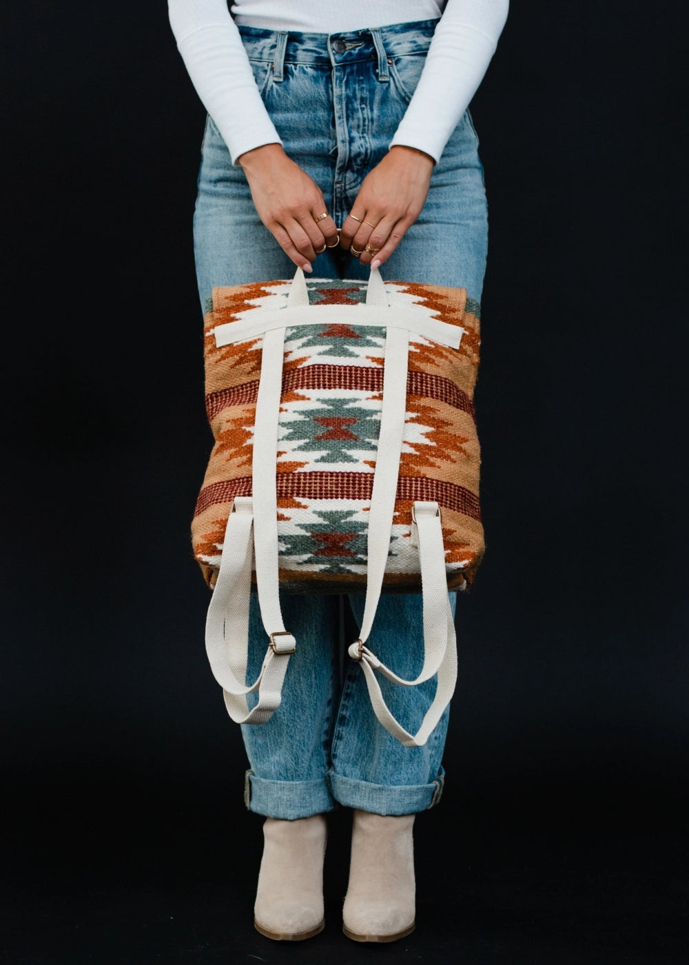 Photograph of a person in light wash cuffed denim jeans and tan suede ankle boots holding a woven backpack with a prominent Southwestern geometric pattern in rust, tan, teal, and cream colors, featuring white canvas straps, against a plain black background