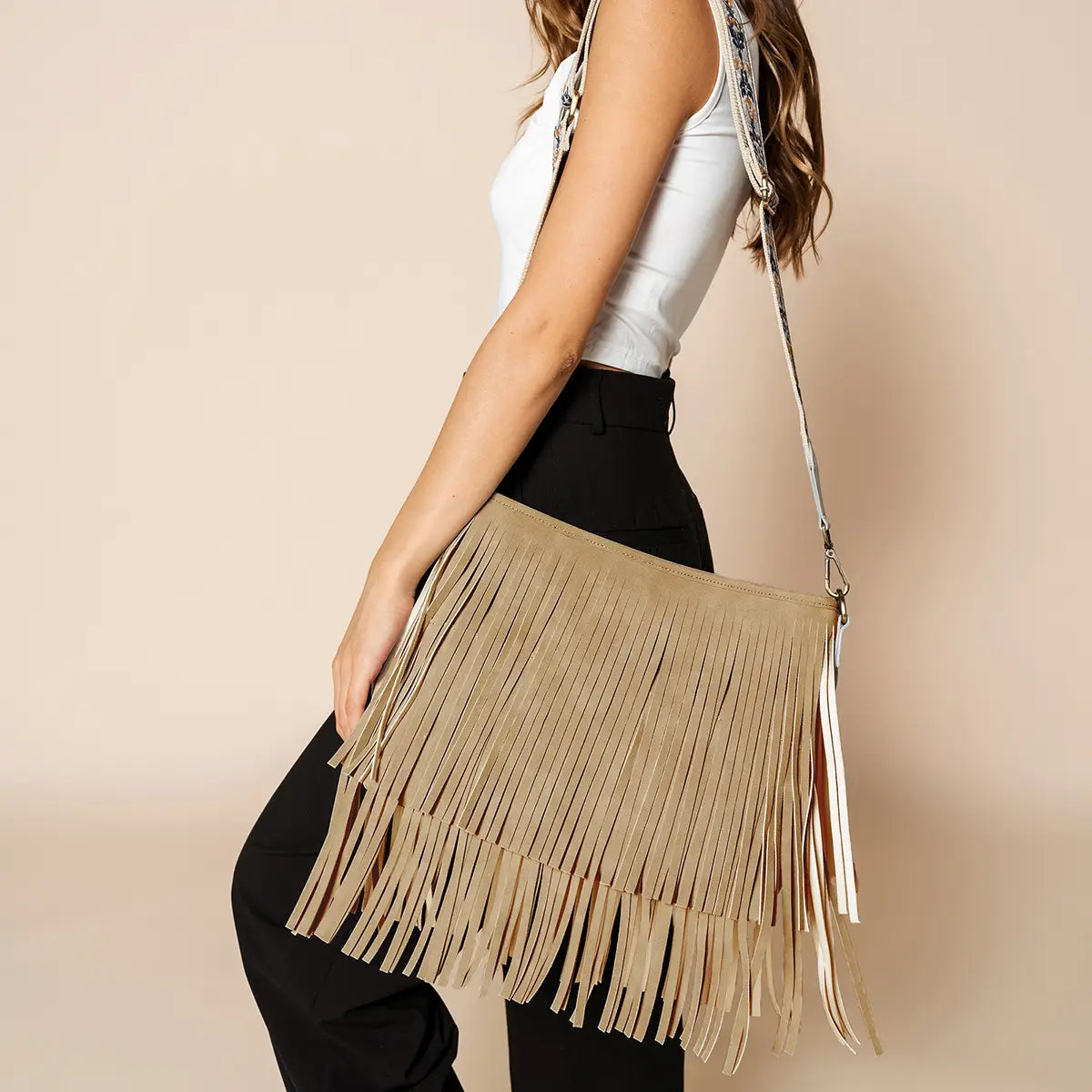 Photography of a fashion model showcasing a large beige suede fringed shoulder bag with a distinct patterned strap, wearing a white tank top and black pants in a minimalist studio setting