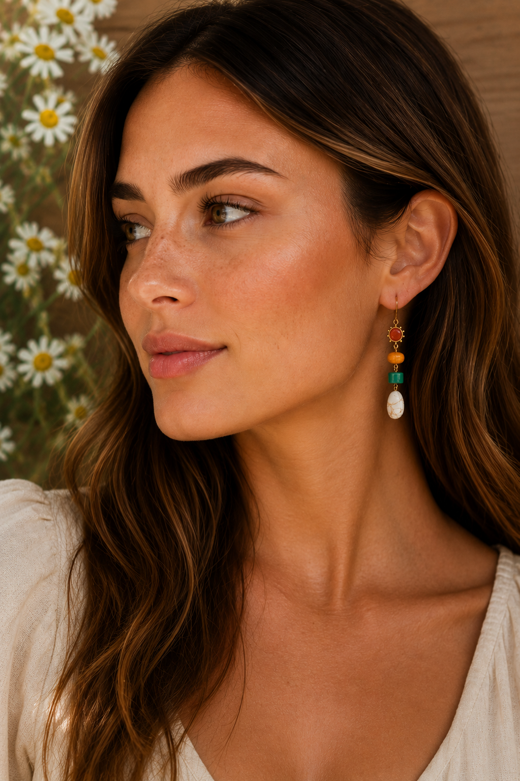 Photograph of a woman's side profile displaying her tanned skin, freckles, and a long colorful beaded earring with red, orange, green, and white stones against a blurred white daisy background