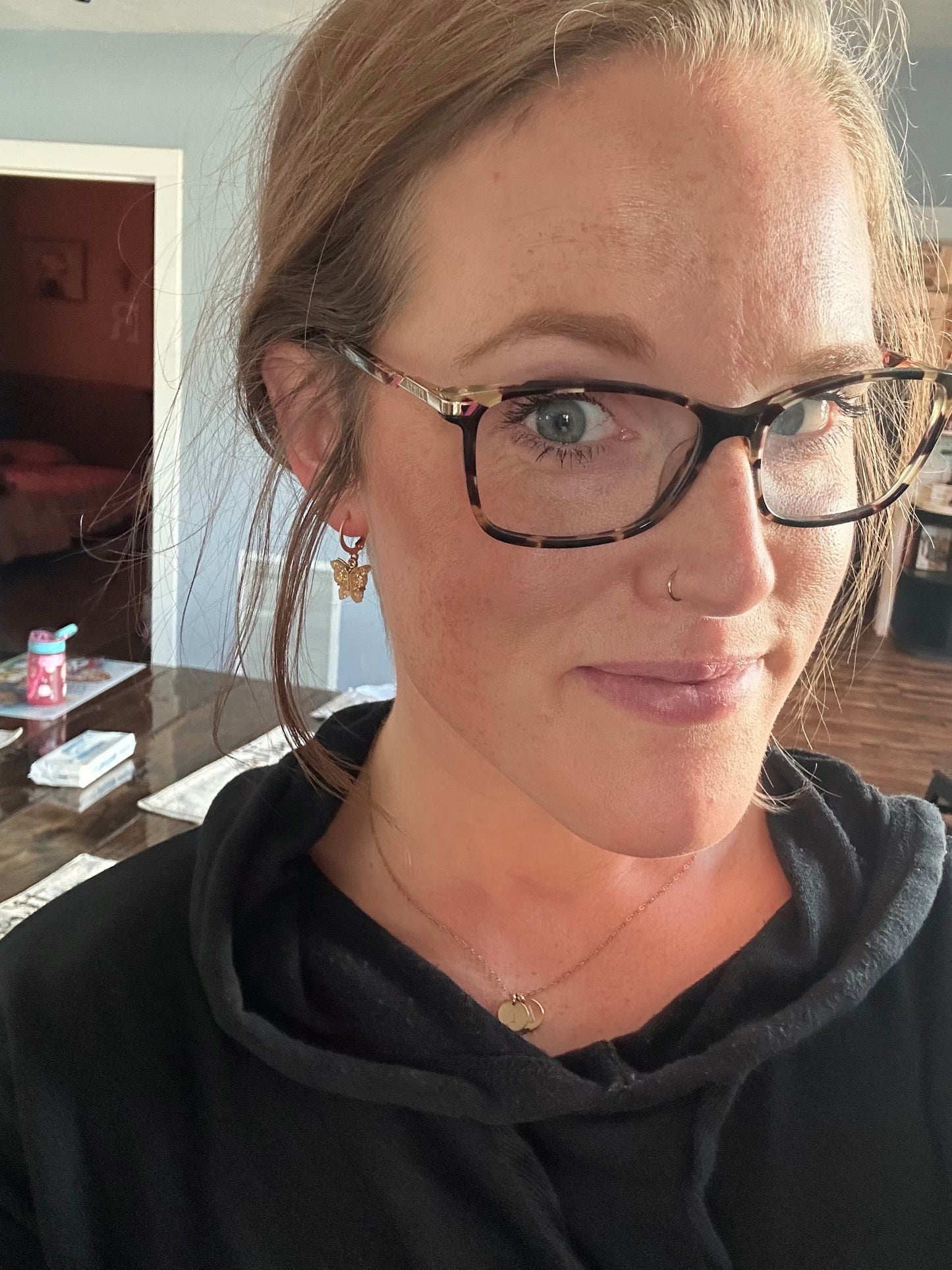 Photograph of a woman with light brown hair and green eyes wearing stylish tortoise-shell glasses a gold butterfly hoop earring and a delicate nose ring in a home setting