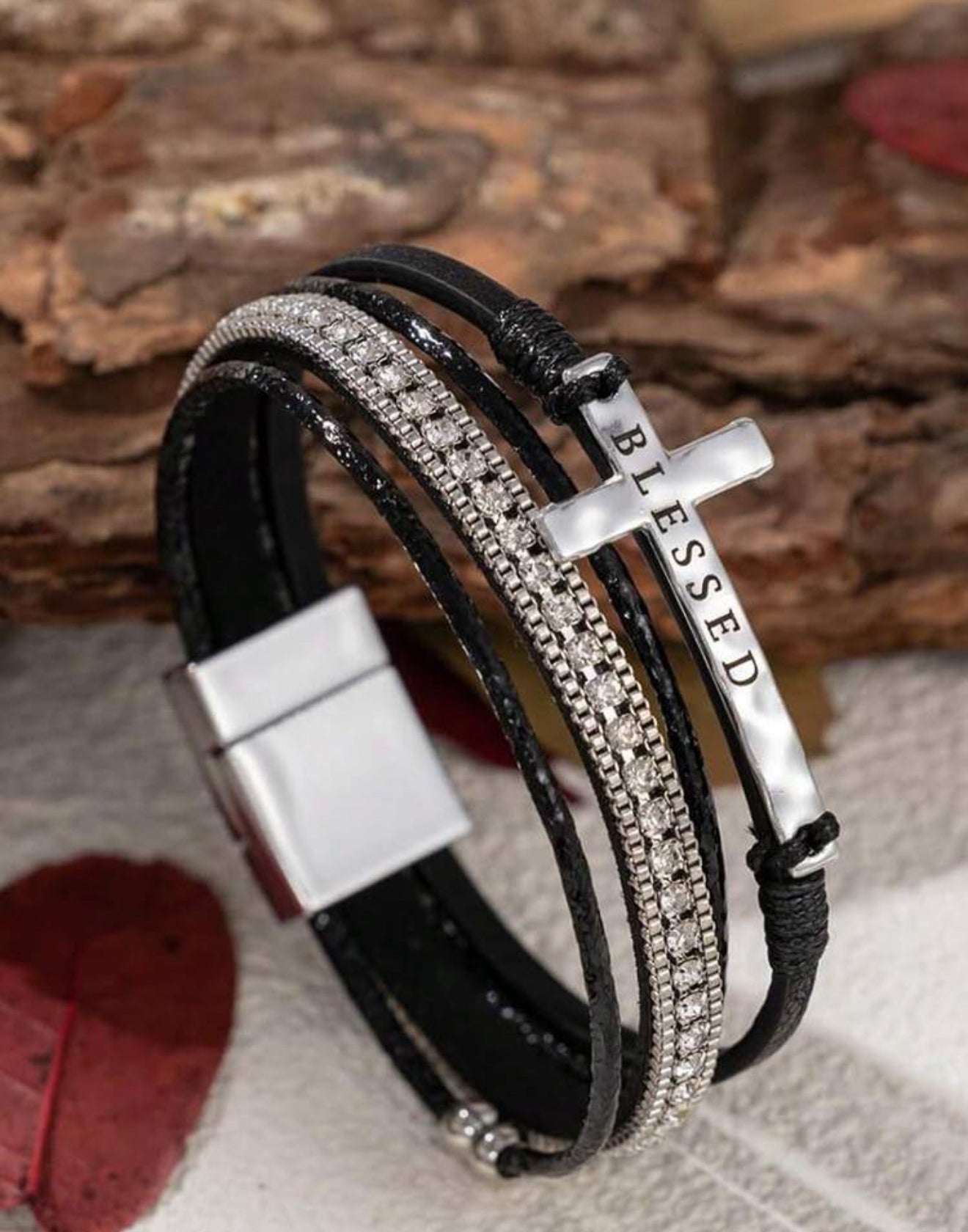 A multi-strand black leather bracelet featuring a silver cross charm engraved BLESSED, a sparkling rhinestone band, and a magnetic clasp against a natural wood and light fabric background