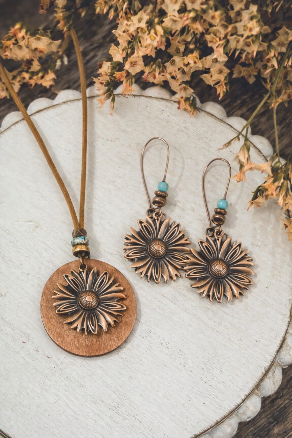 A bohemian-style copper-toned daisy motif jewelry set including a necklace with a brown leather cord and matching drop earrings featuring small turquoise beads, displayed on a rustic white scalloped wooden surface with dried light brown flowers in the background