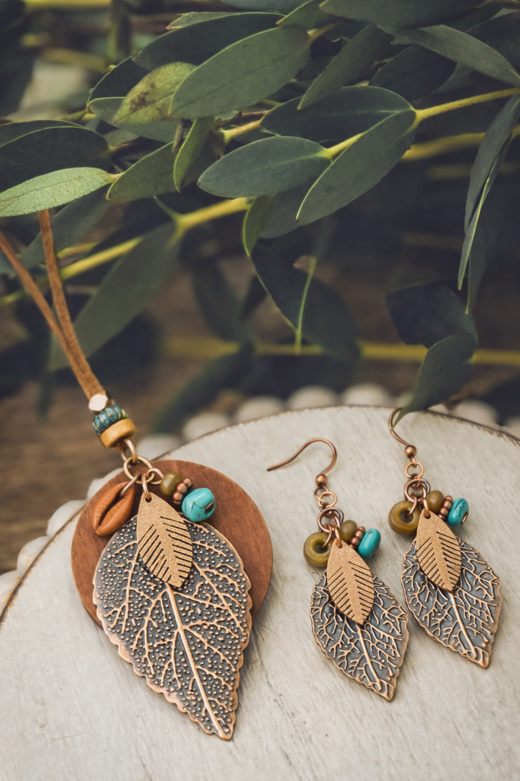 Photograph of a bohemian copper leaf necklace featuring intricate veining, a cowrie shell, and turquoise beads on a brown suede cord, displayed with matching copper leaf dangle earrings adorned with turquoise and olive beads, against a backdrop of blurred green eucalyptus leaves