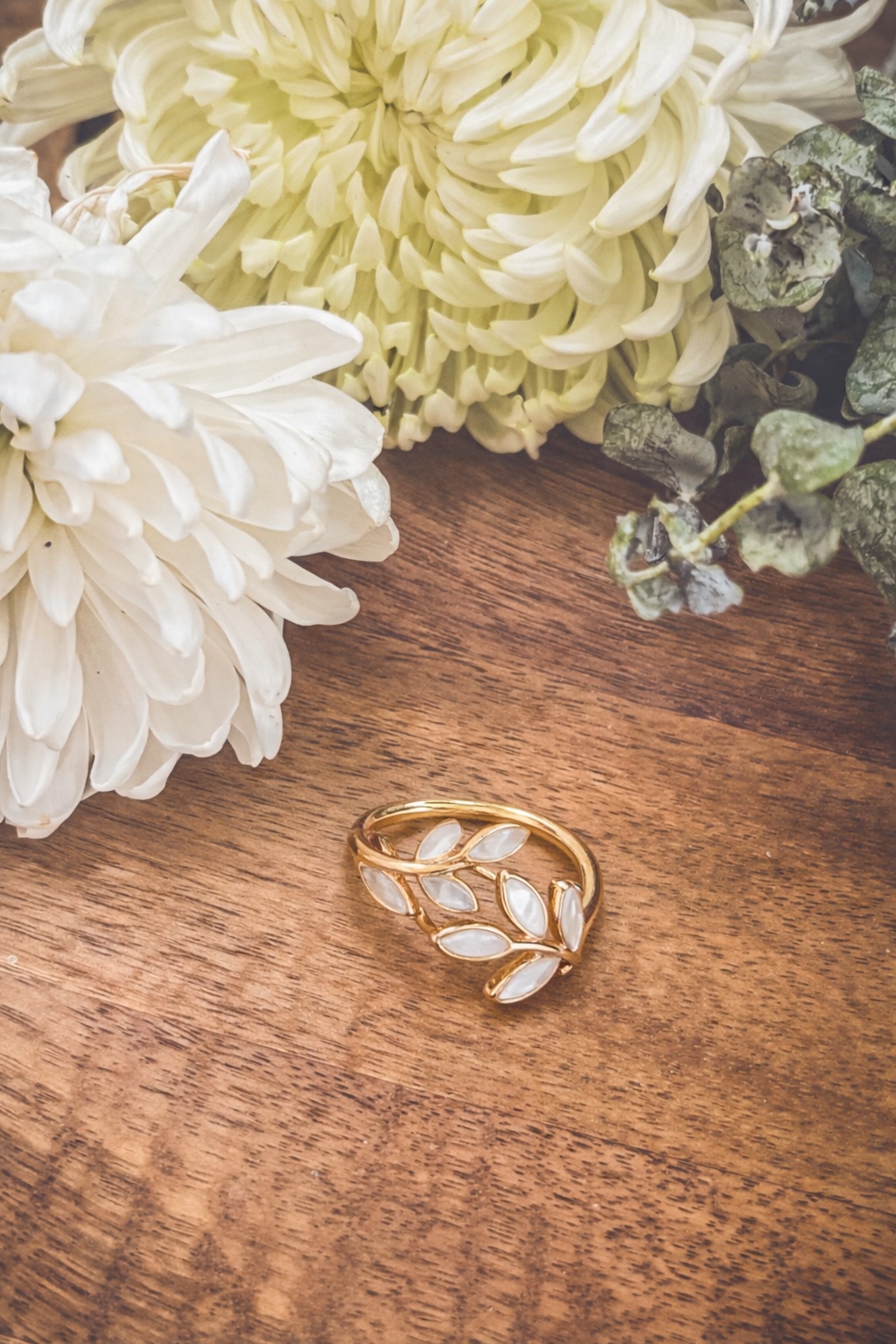 Gold ring featuring a mother-of-pearl leaf design resting on a rustic wooden surface with fluffy white and light green chrysanthemum blossoms and subtle eucalyptus foliage framing the top