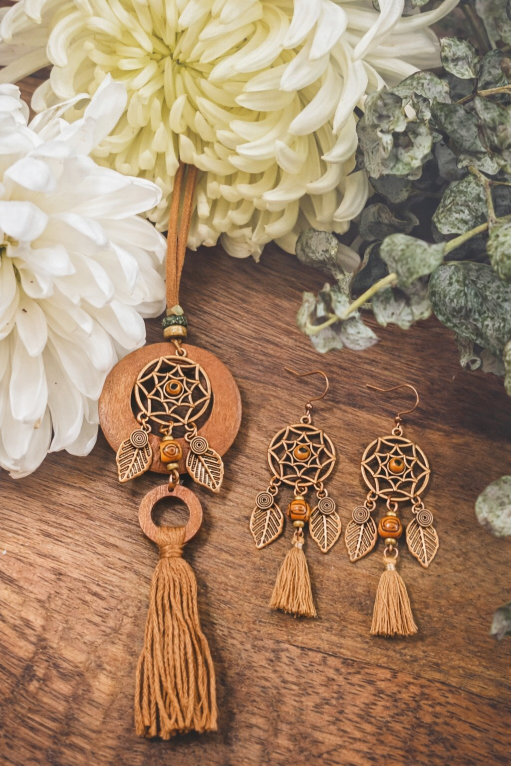 Bohemian wooden and bronze dreamcatcher necklace with tassel and matching dangle earrings displayed on rustic wood next to creamy white chrysanthemums and muted green foliage