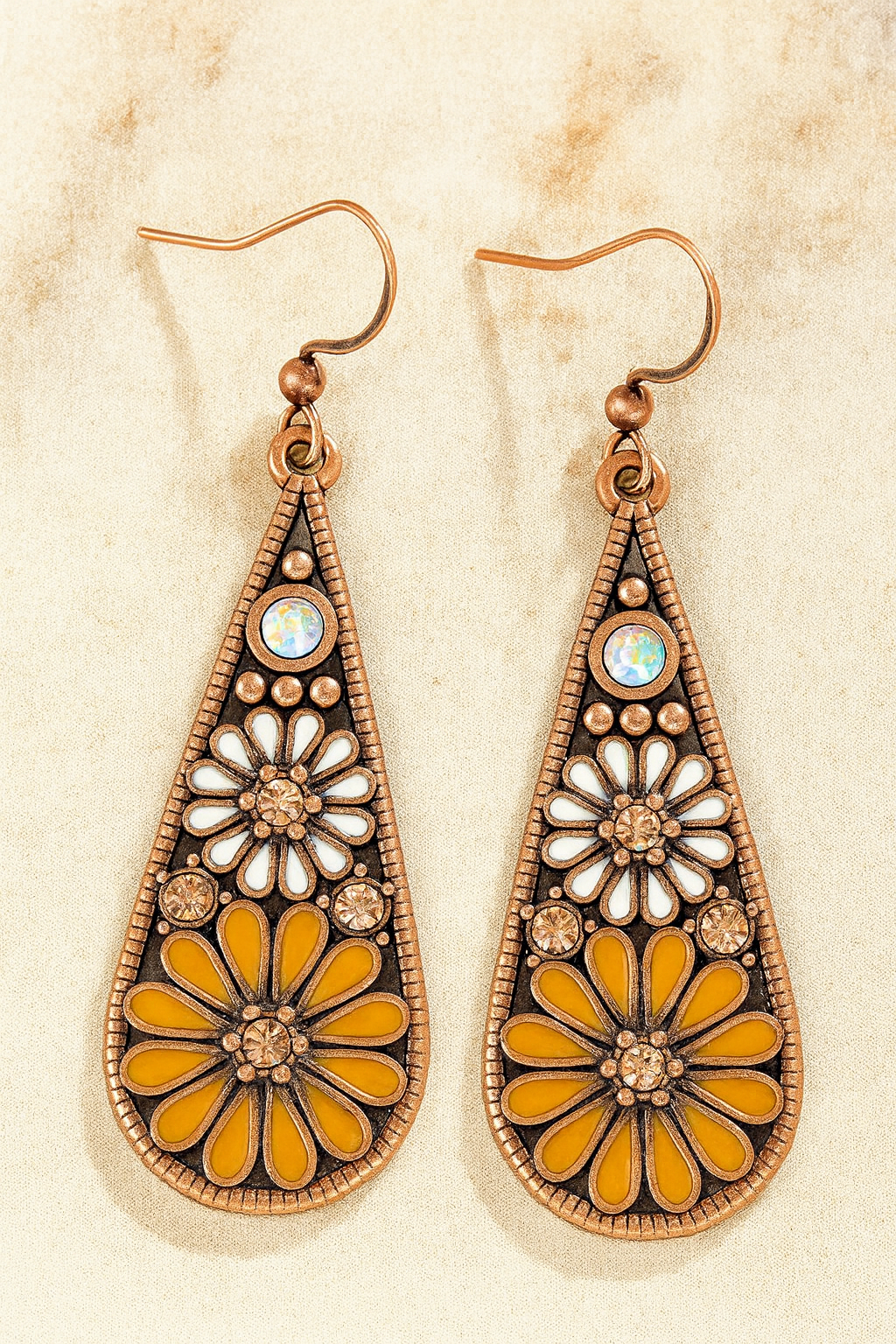 A pair of copper-toned dangle earrings on a light fabric surface featuring teardrop pendants with white and mustard yellow enamel floral patterns an iridescent top stone and clear rhinestone accents