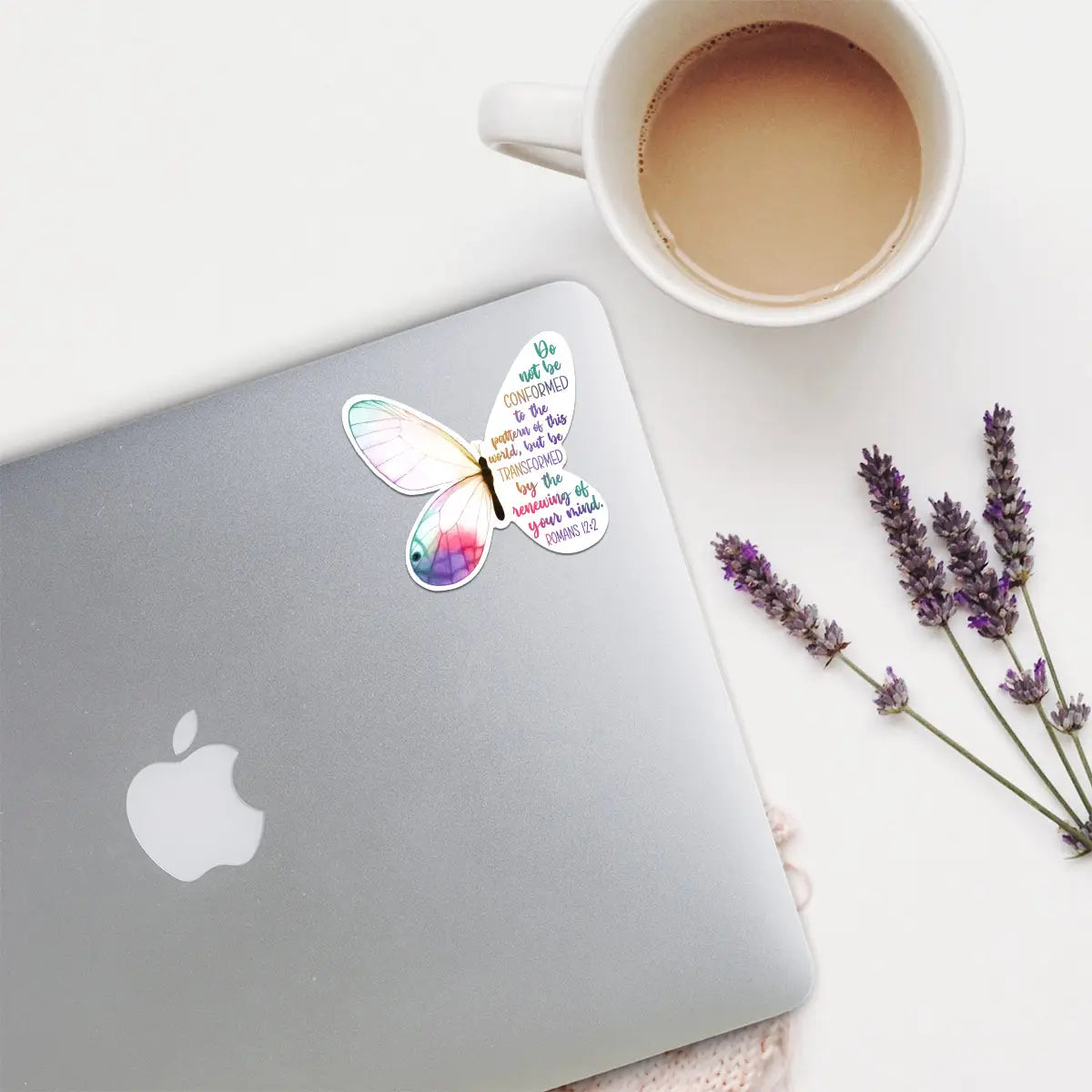 A colorful watercolor-style butterfly sticker with the quote "Do not be conformed to the pattern of this world, but be transformed by the renewing of your mind. ROMANS 12:2" adhered to a silver Apple MacBook laptop, displayed next to a white mug of coffee and three sprigs of dried purple lavender flowers on a bright white tabletop
