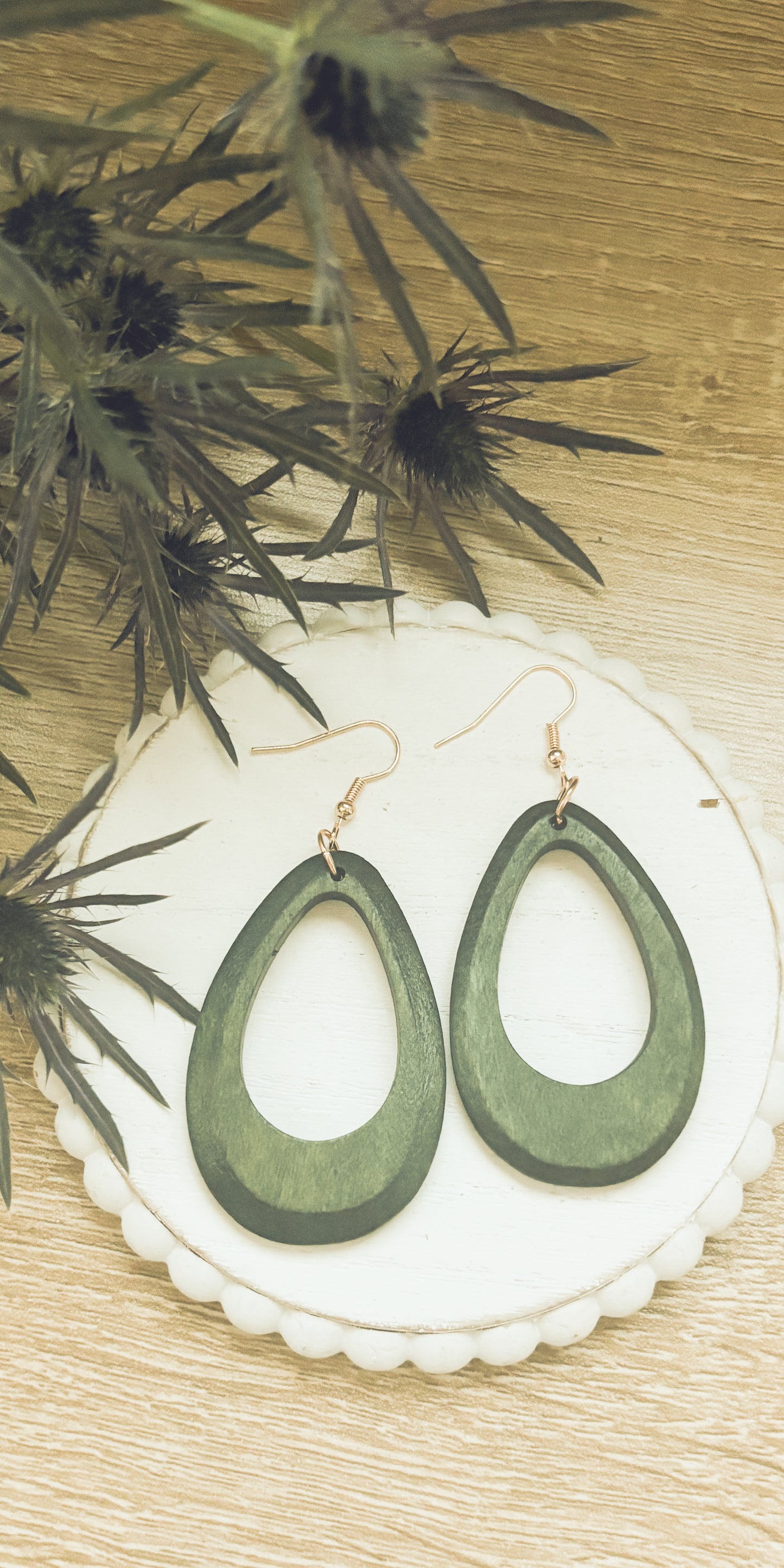 A pair of dark olive green wooden teardrop earrings with rose gold hooks rests on a white scalloped coaster next to a blurry spiky green thistle plant on a light wood table