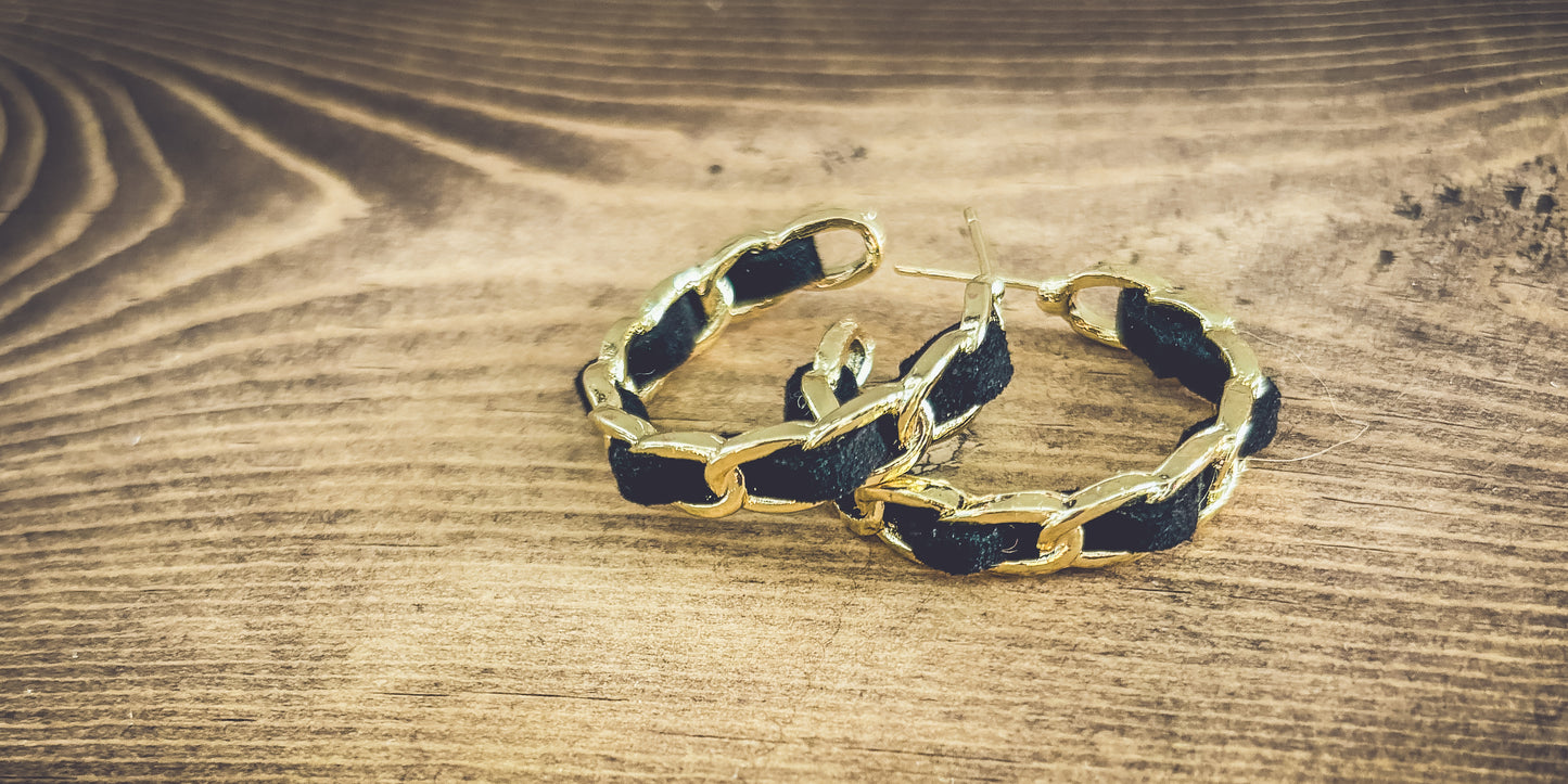 A pair of gold chain link hoop earrings intricately woven with black fabric resting on a rustic light brown wooden surface