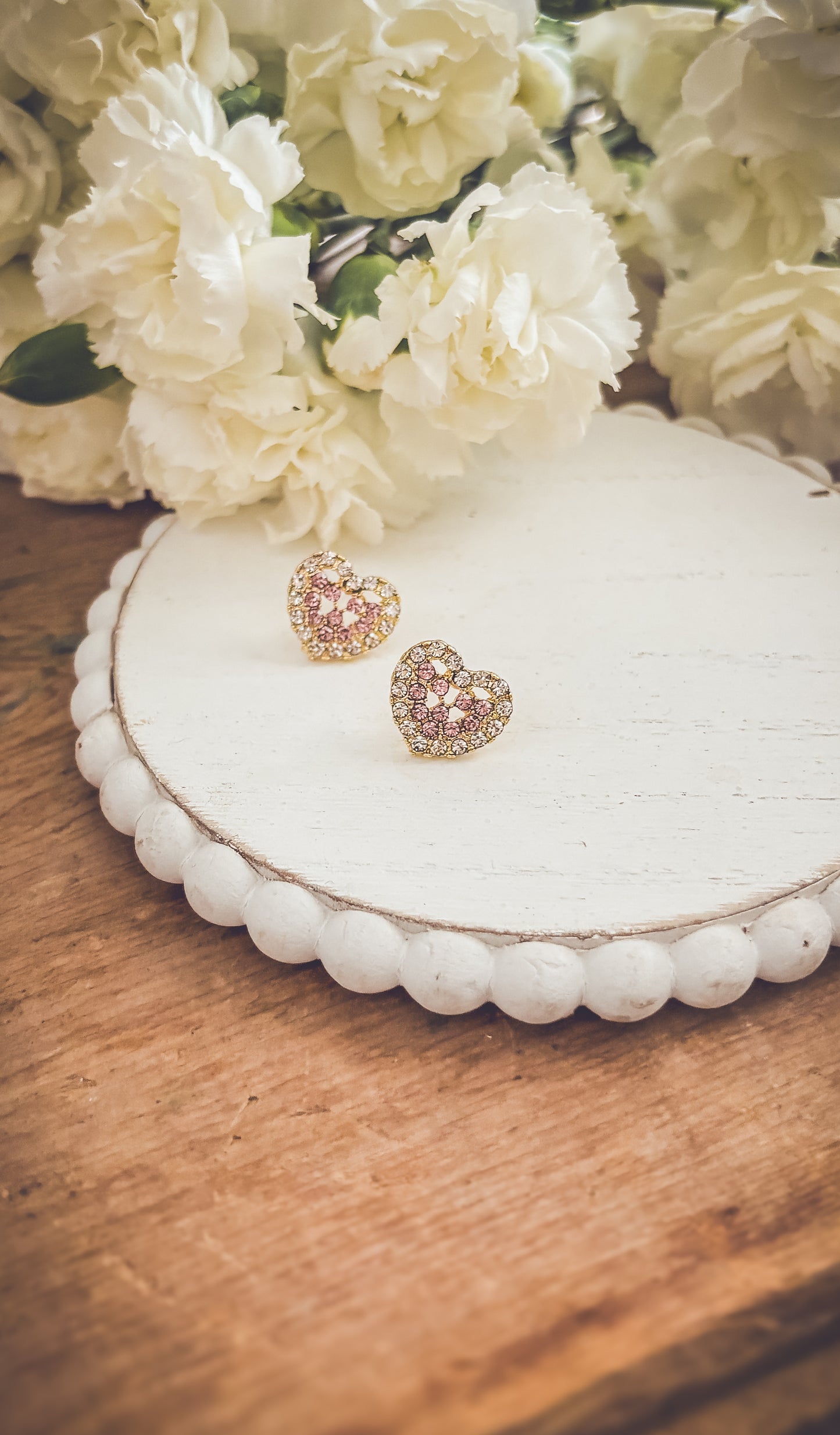 Photograph of two sparkling gold heart-shaped earrings adorned with pink and clear gemstones resting on a rustic white scallop-edged wooden stand with soft white carnations blooming in the background