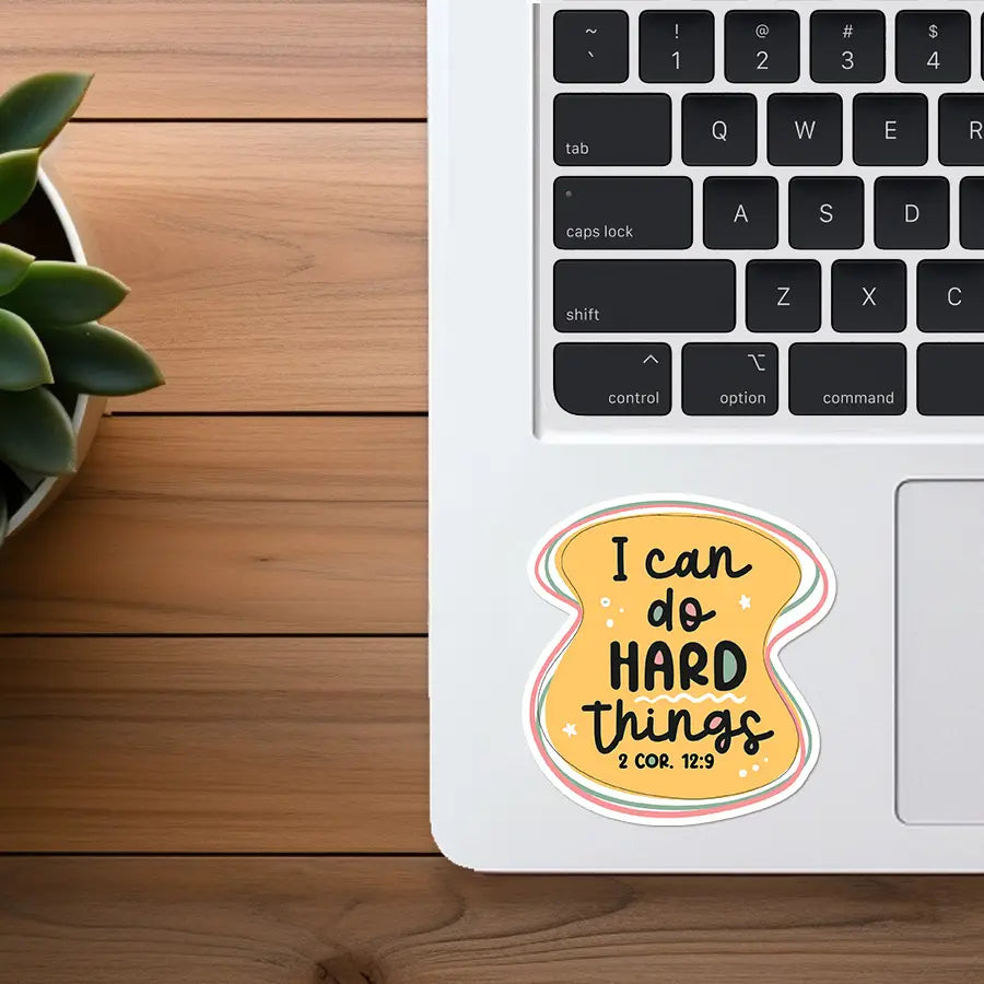 Digital image of a yellow irregular-shaped motivational sticker with black text reading I can do HARD things 2 COR. 12:9, outlined in green and pink, adorned with tiny stars and dots, placed on a white laptop keyboard area alongside a wooden desk and a green potted succulent plant