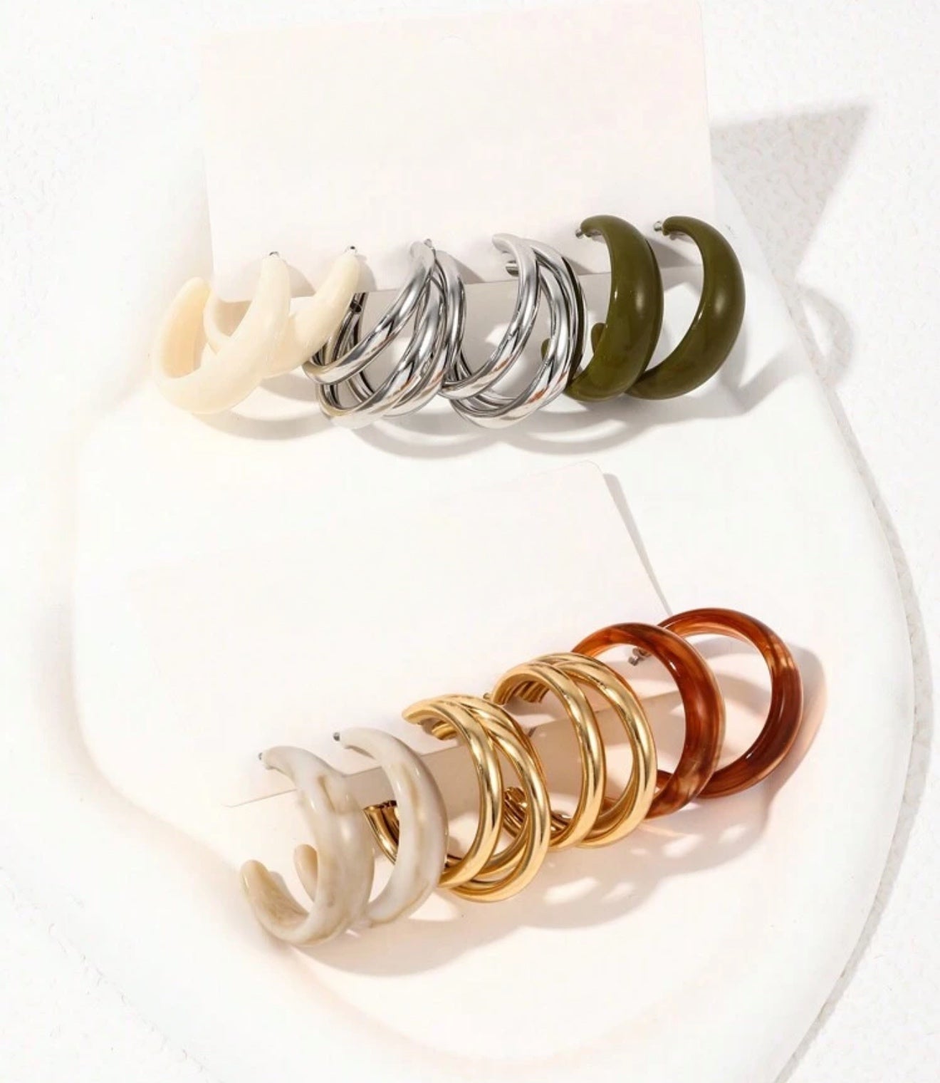 A curated collection of contemporary hoop earrings displayed on a white stand showcasing chunky cream, silver triple, olive green, marble beige, gold triple, and amber brown styles