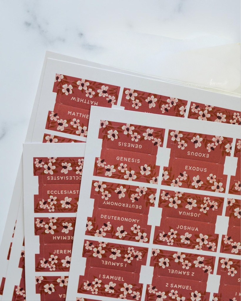 Terracotta Bible book tab sticker sheets with white and brown floral garland patterns and book names like Genesis and Matthew fanned on a light surface