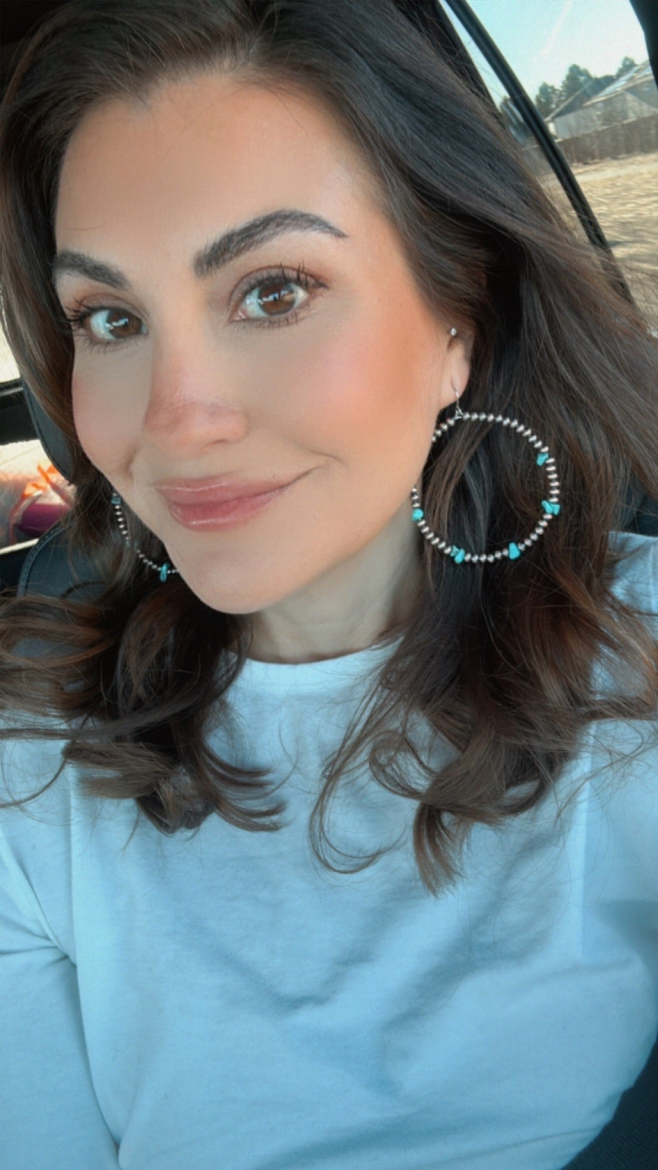 A smiling woman with long brown hair, brown eyes, and light pink blush wearing a white t-shirt and turquoise and silver beaded hoop earrings taking a selfie inside a car with a blurred outdoor background