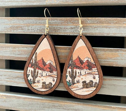 A pair of wooden teardrop earrings displaying a printed desert landscape illustration featuring tall saguaro cacti, red rock mountains, and an orange sunset, accented with dark wood frames and gold-colored hooks