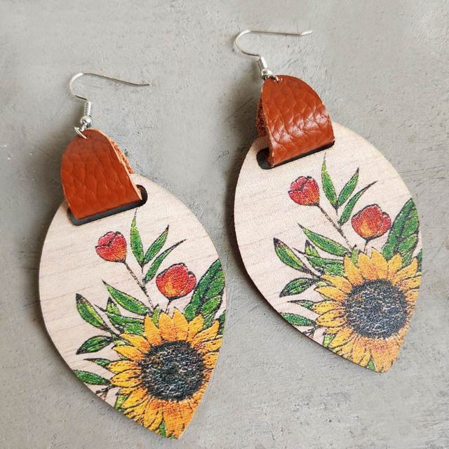 Pair of wooden leaf-shaped dangle earrings with brown faux leather accents featuring a colorful floral print of yellow sunflowers and red tulips on a grey surface