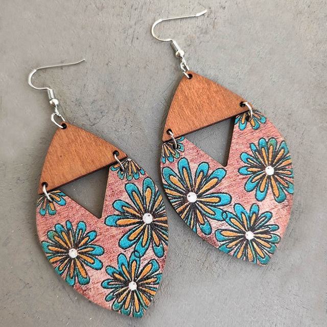Wood and metal teardrop dangle earrings with a split design featuring a natural brown wood top and a lower faded pinkish section decorated with light blue and orange daisy-like flowers having white centers, displayed on a grey surface