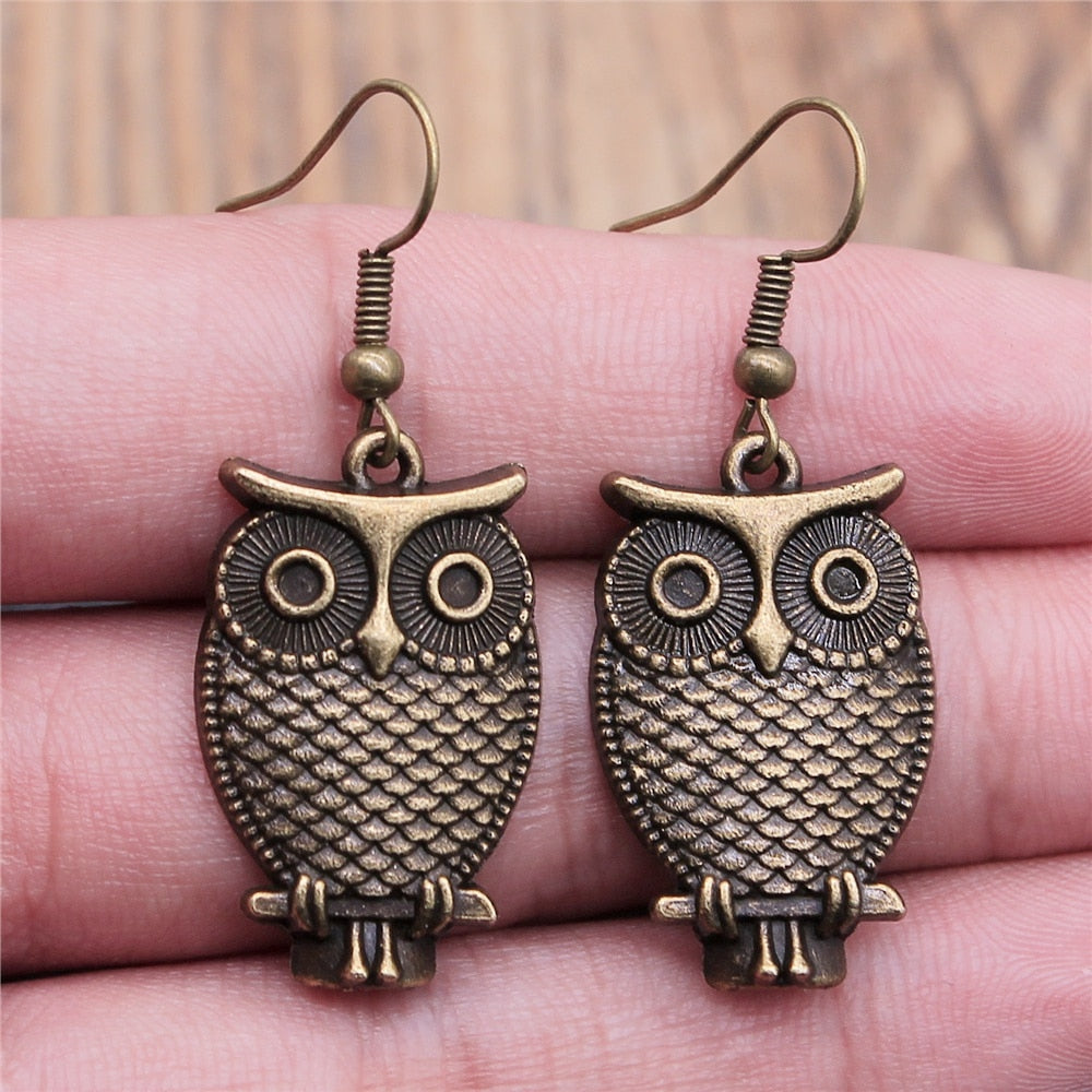 Pair of antique bronze owl dangle earrings held in a hand featuring intricate feather details and prominent circular eyes with a rustic finish
