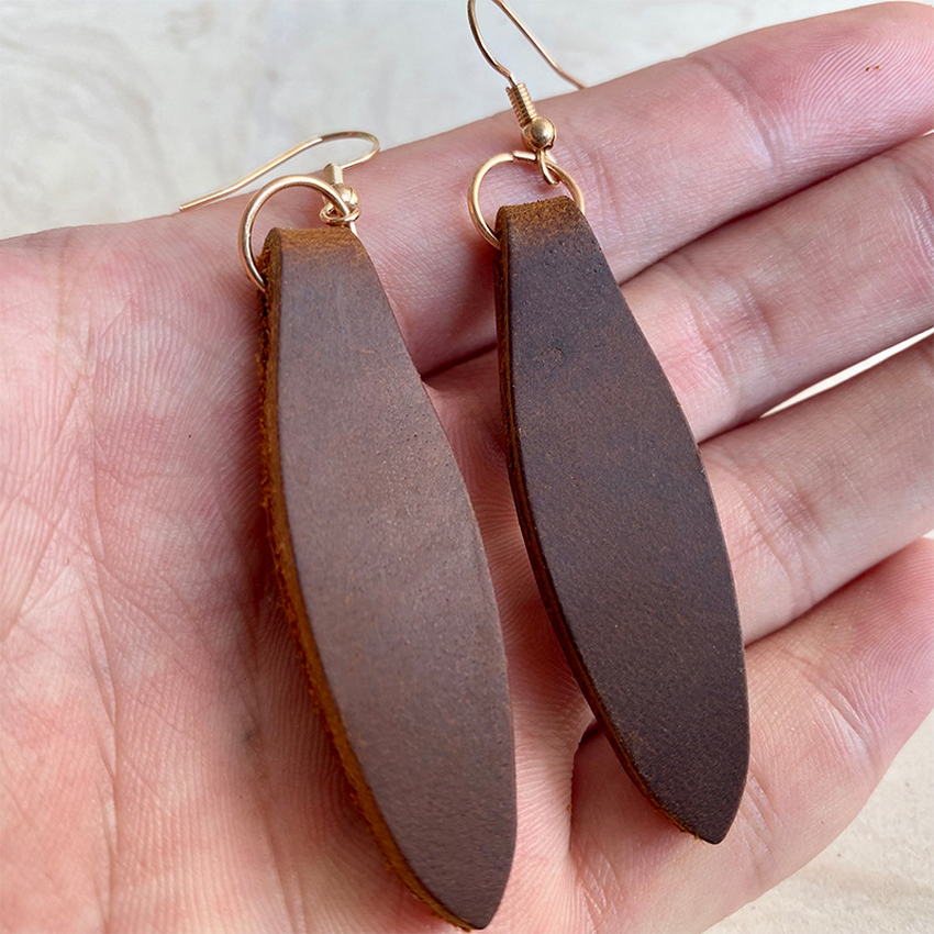 Handmade dark brown leather earrings with an elongated leaf shape and gold-toned fishhook findings displayed in a hand