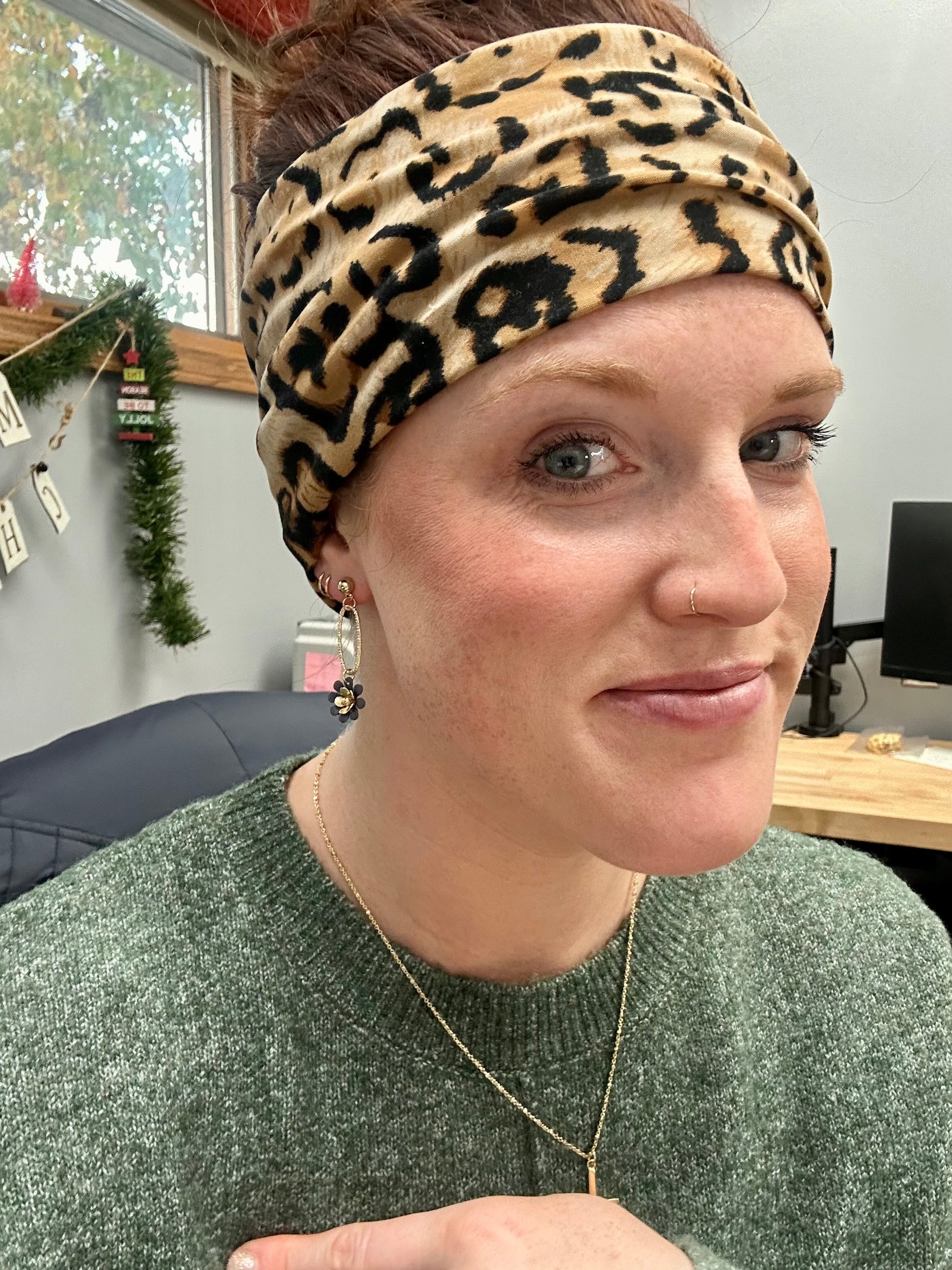 Photograph of a smiling woman in an office setting wearing a leopard print headband a green knit sweater and a festive Christmas garland visible in the background