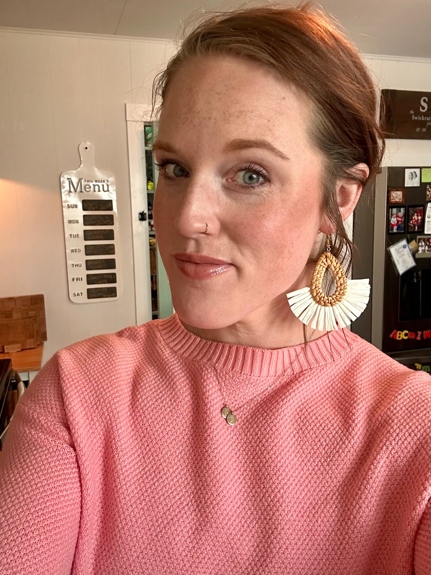 Selfie of a woman with auburn hair and blue eyes wearing a pink textured sweater and distinctive fan-shaped raffia earrings in a home kitchen featuring a "This Week's Menu" board