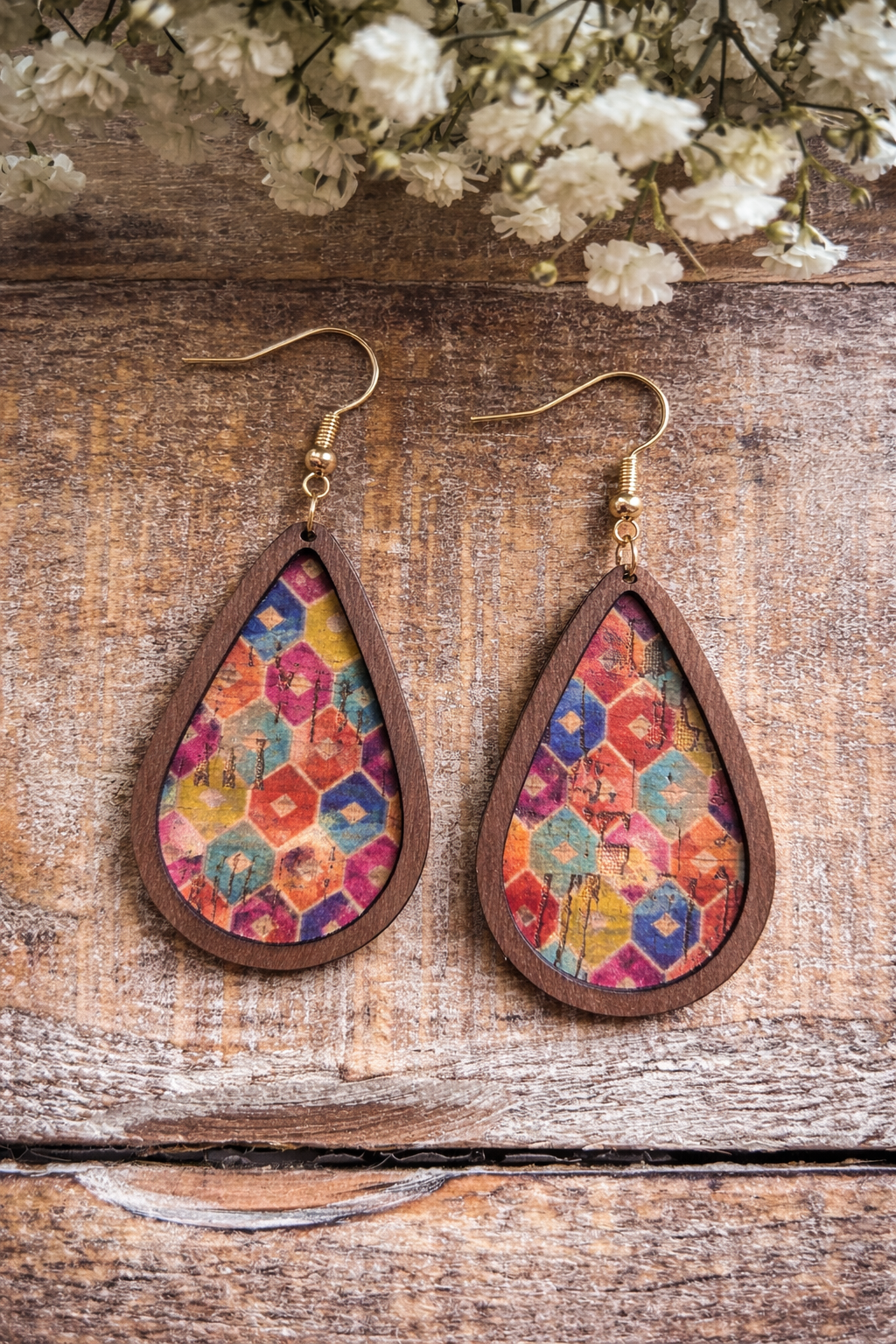 A pair of handcrafted teardrop wooden earrings featuring colorful hexagonal patterned cork inserts and golden hooks rests on a rustic wooden surface accented by delicate white baby's breath flowers