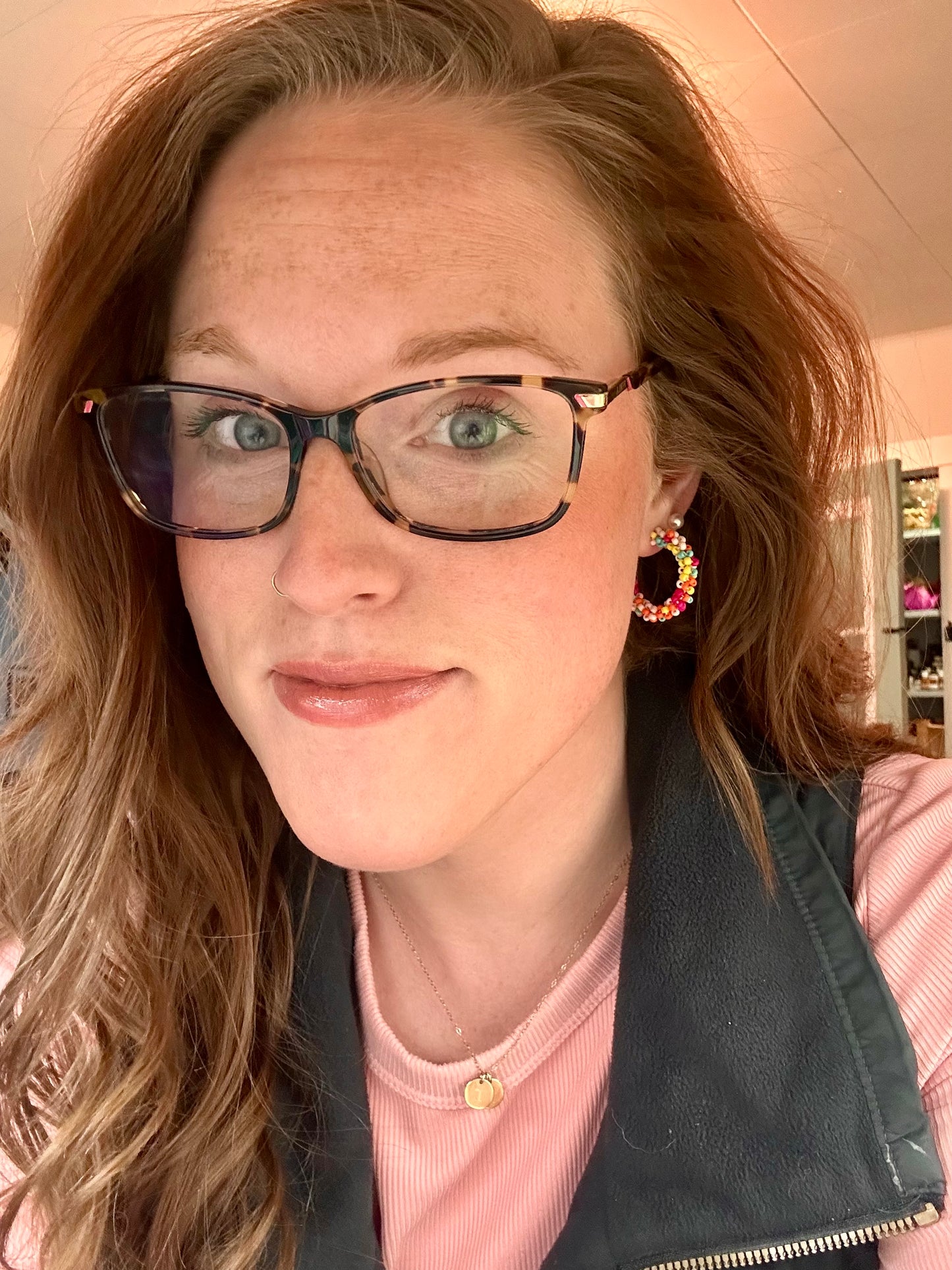 Headshot of a woman with auburn hair and green eyes, wearing tortoiseshell glasses, a gold nose ring, and vibrant multicolor beaded hoop earrings, against an indoor background