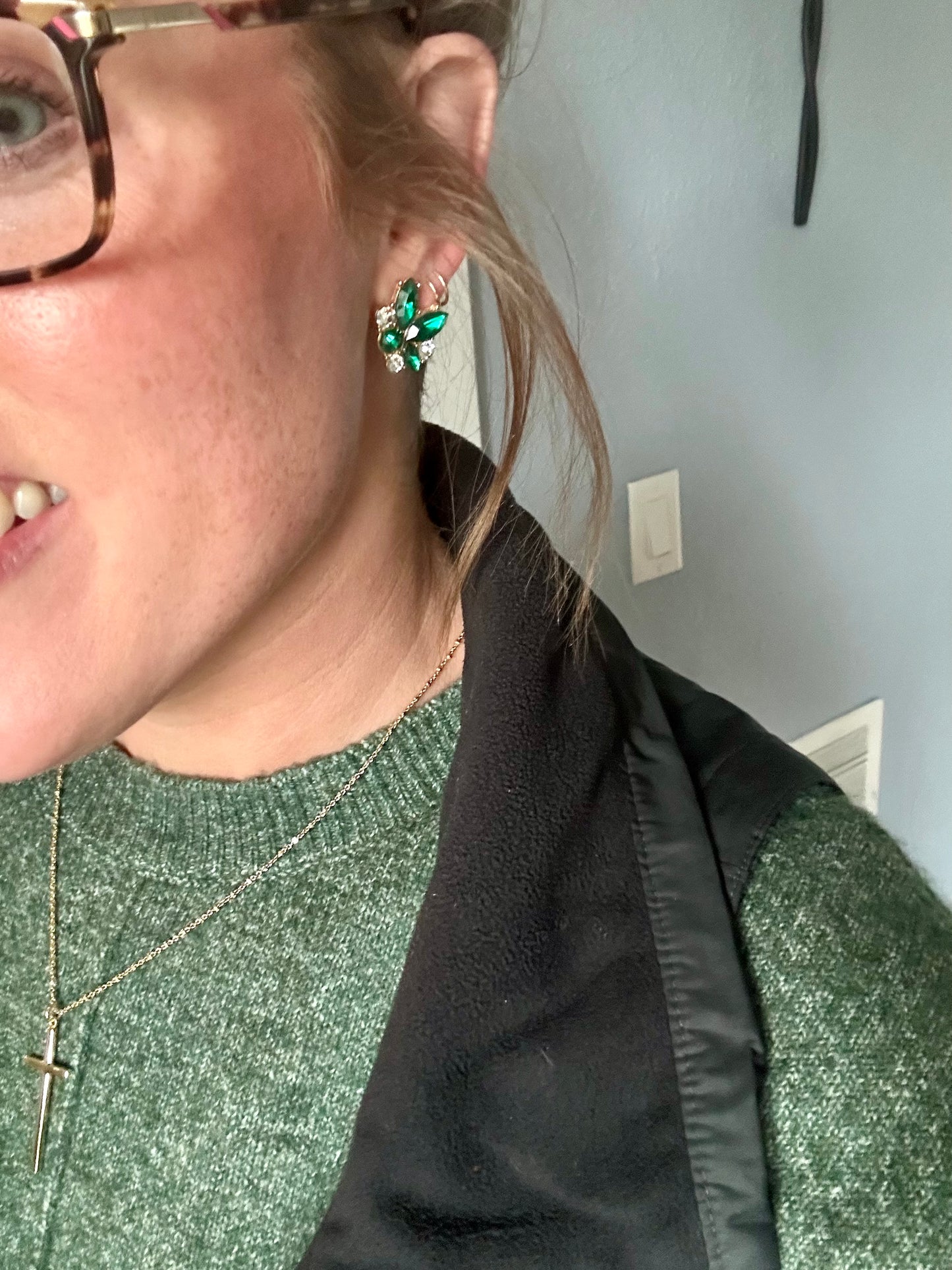 Close-up of a person indoors wearing a green ornate stud earring a gold cross necklace and a green knit sweater