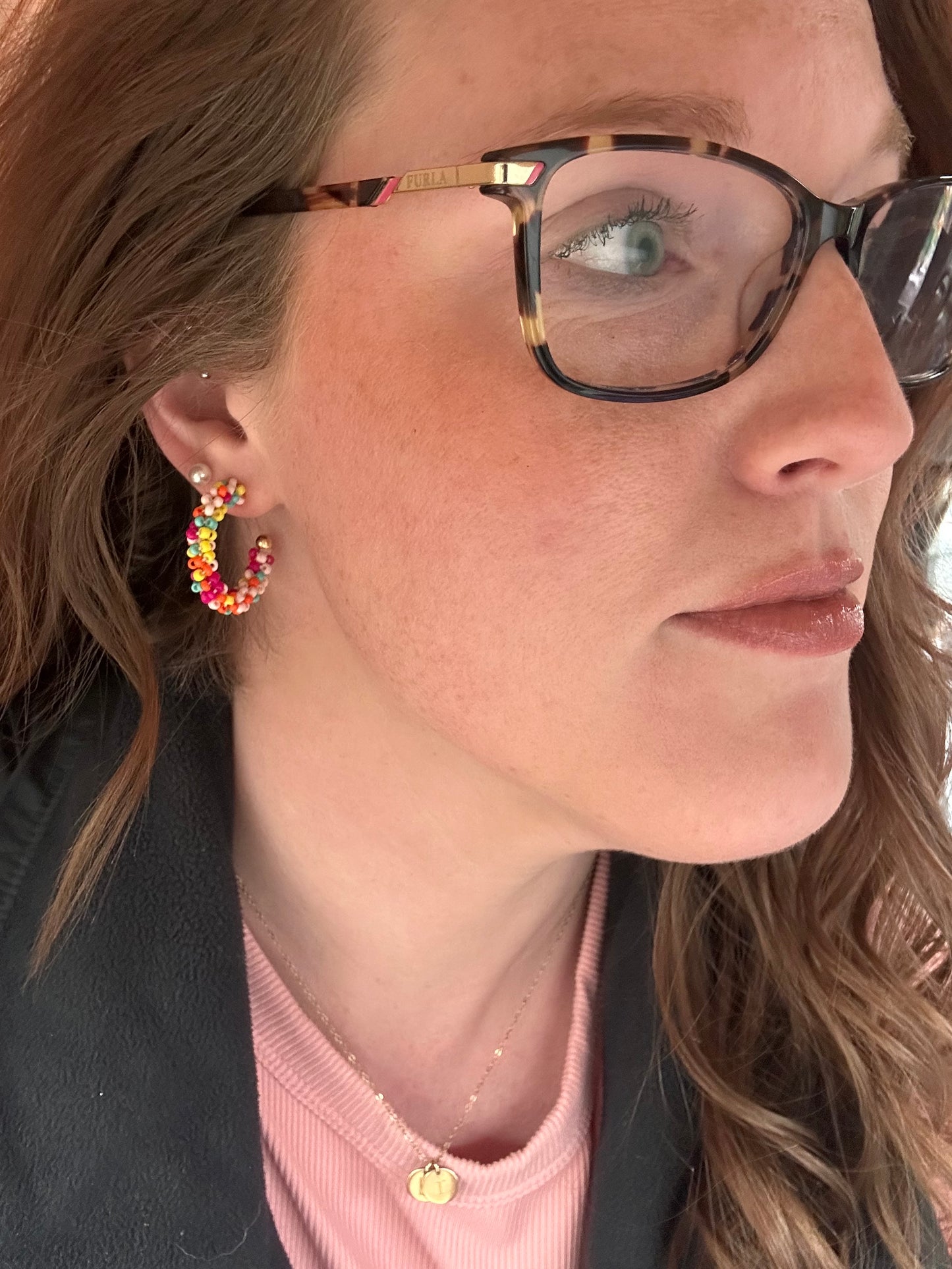 Photograph of a woman's side profile showcasing tortoiseshell Furla eyeglasses a vibrant multi-colored beaded hoop earring and a delicate gold initial necklace