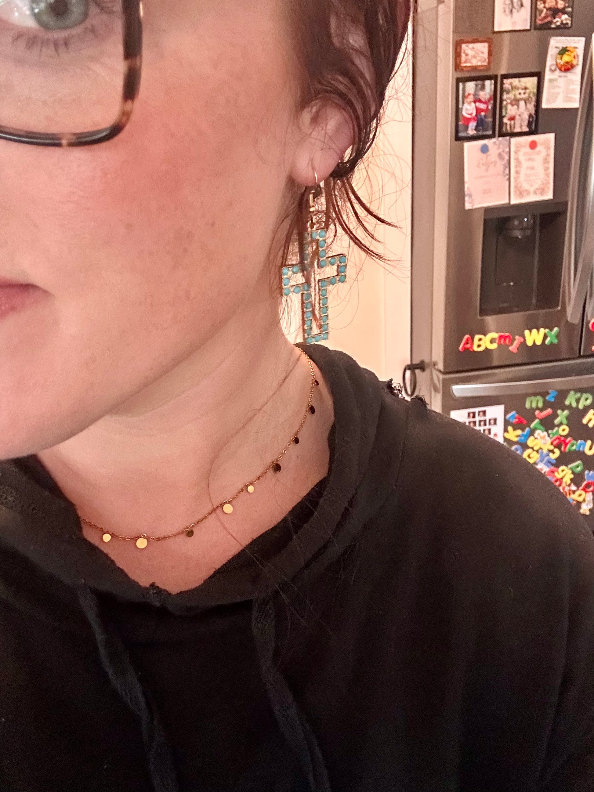 Close-up of a person wearing a large gold and turquoise cross earring and a gold disc choker necklace, standing in a kitchen with a refrigerator displaying magnetic alphabet letters and family photos