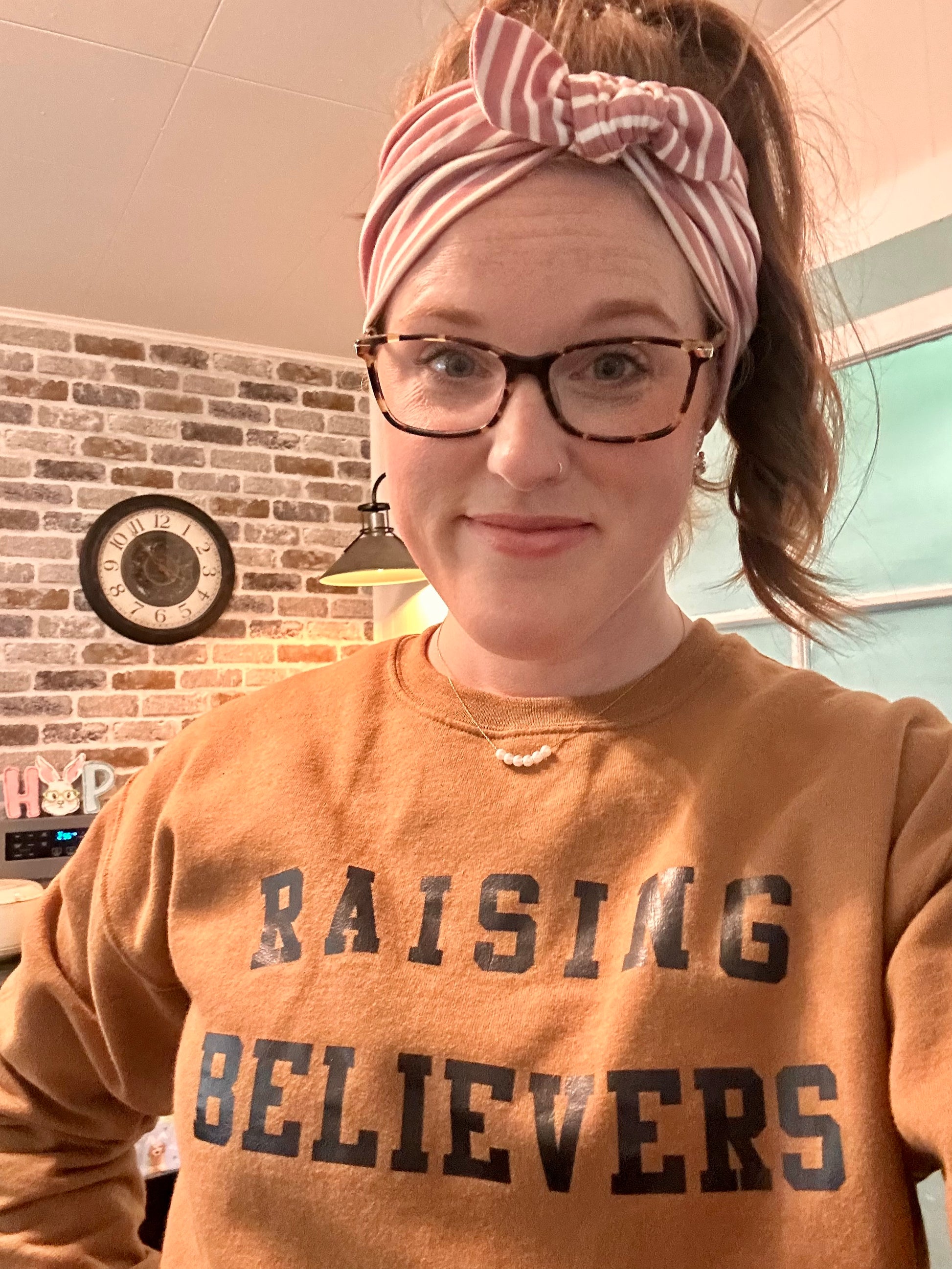 Smiling woman with red hair and a pink striped headband wearing tortoiseshell glasses, a rust-colored 'RAISING BELIEVERS' sweatshirt, a pearl necklace, and an industrial lamp earring, posing indoors against a brick wall with a vintage clock