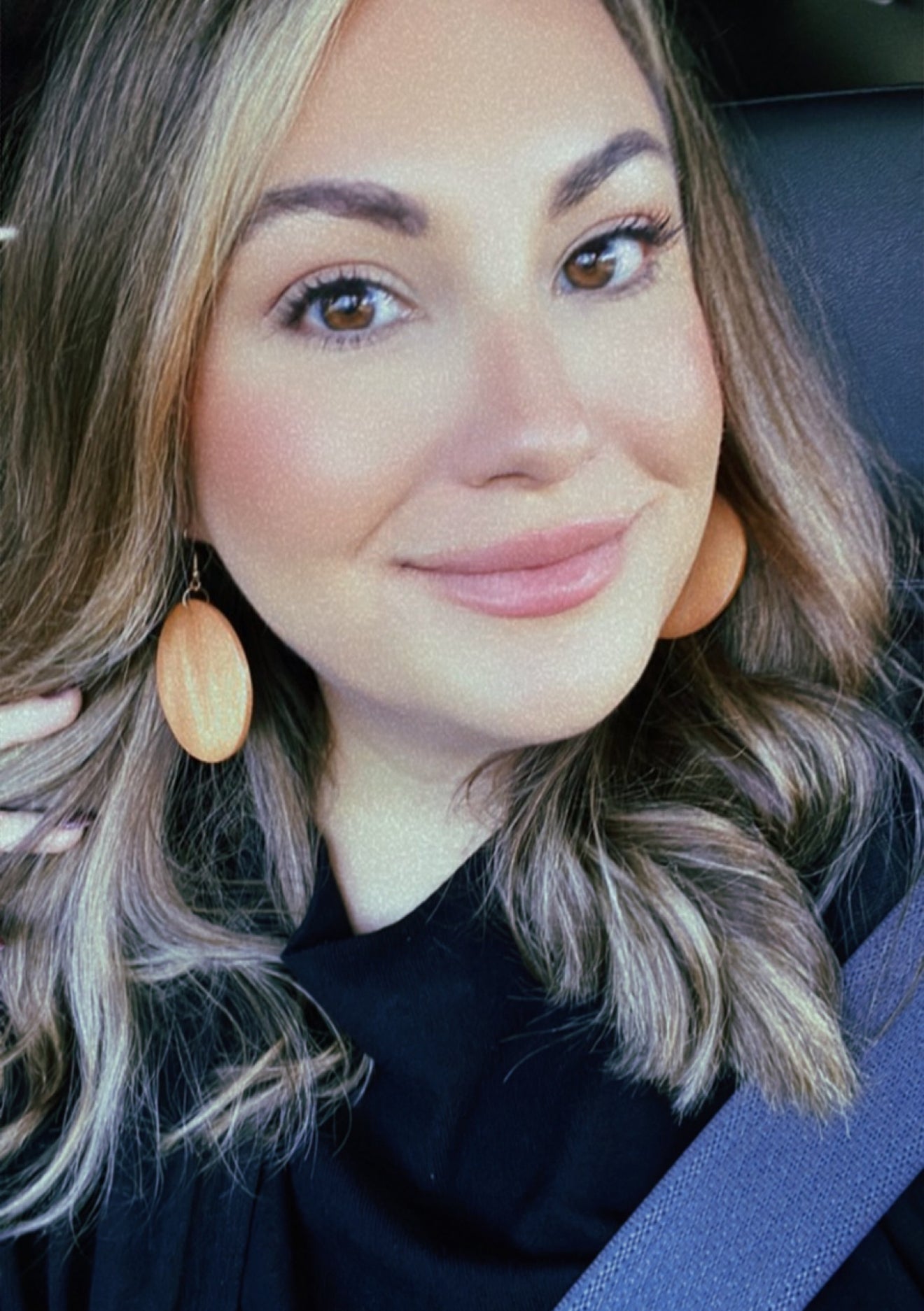A smiling woman with wavy blonde and brown hair wears wood-toned oval earrings and a blue seatbelt in a car